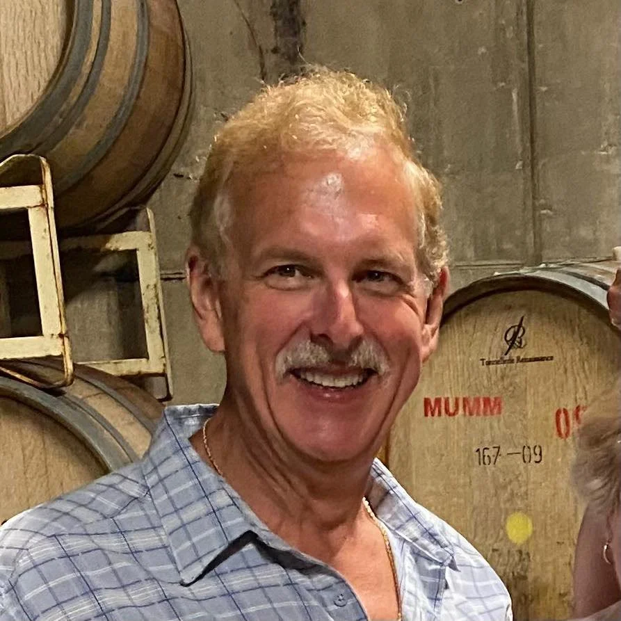 A smiling man with light-colored hair and a mustache in front of wine barrels in a cellar.