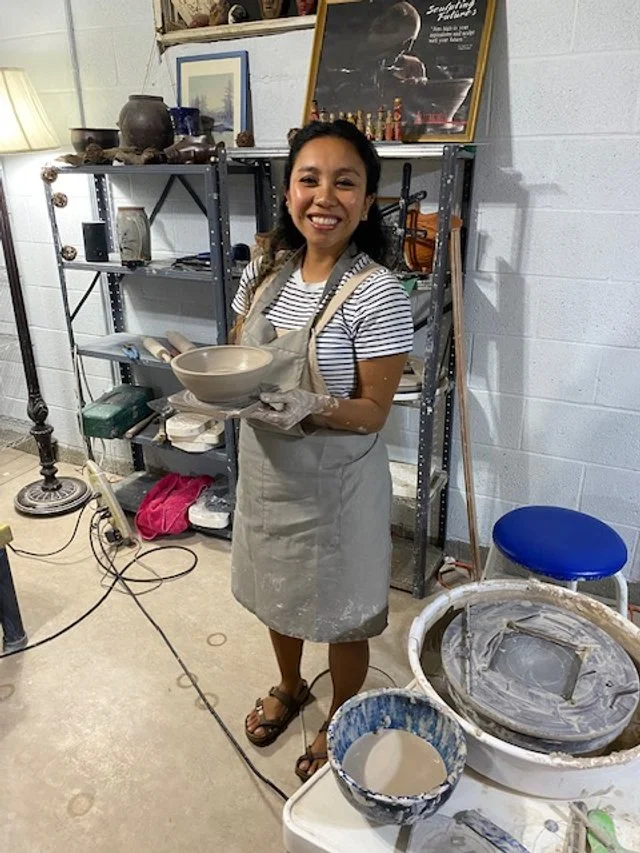 A woman smiling while holding a ceramic bowl, standing in a pottery studio with shelves of pottery and art on the wall behind her.