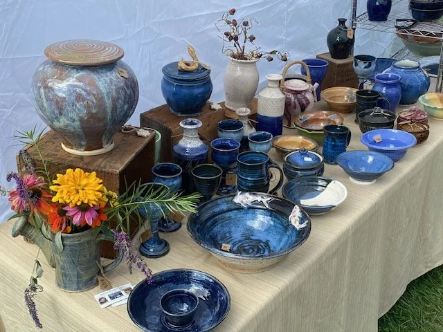 Display of various pottery and ceramic items on a table, including vases, bowls, and jars in blue, earthy, and pastel colors, with some flowers in a pitcher on the side.
