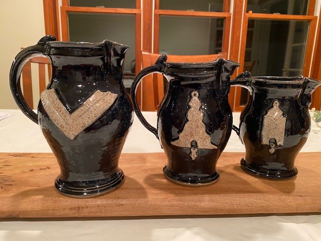 Three black ceramic pitchers with brown patterns, arranged on a wooden board on a table.