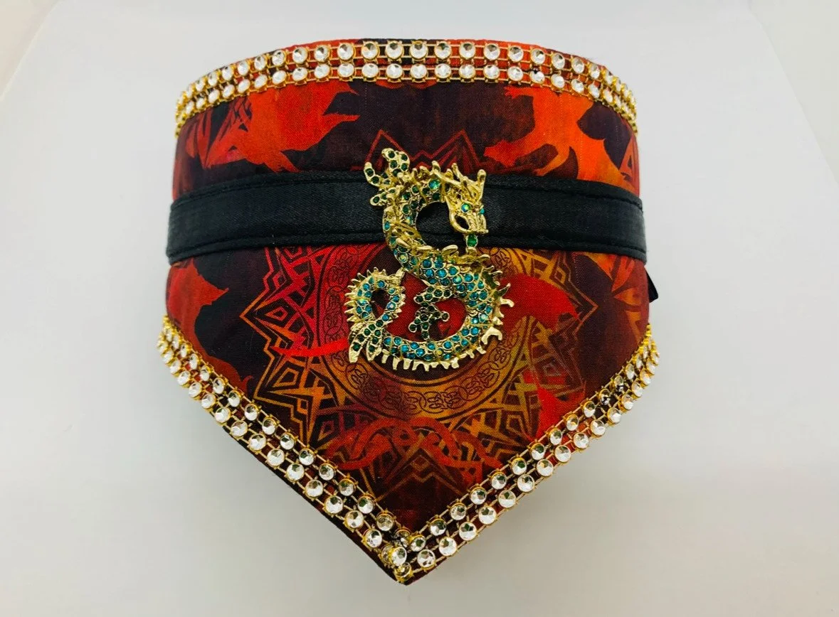 A decorative box with a red and black patterned fabric covering, trimmed with rhinestones along the edges, and a black band around the middle. A gold dragon-shaped brooch with blue and green gemstones is attached to the black band.
