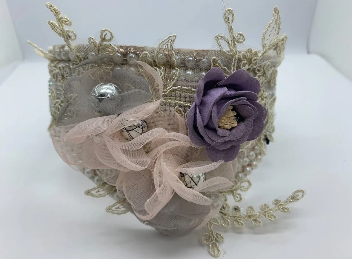 Decorative fabric headband with lace embroidery, pearls, and fabric flowers in purple, pink, and gray.