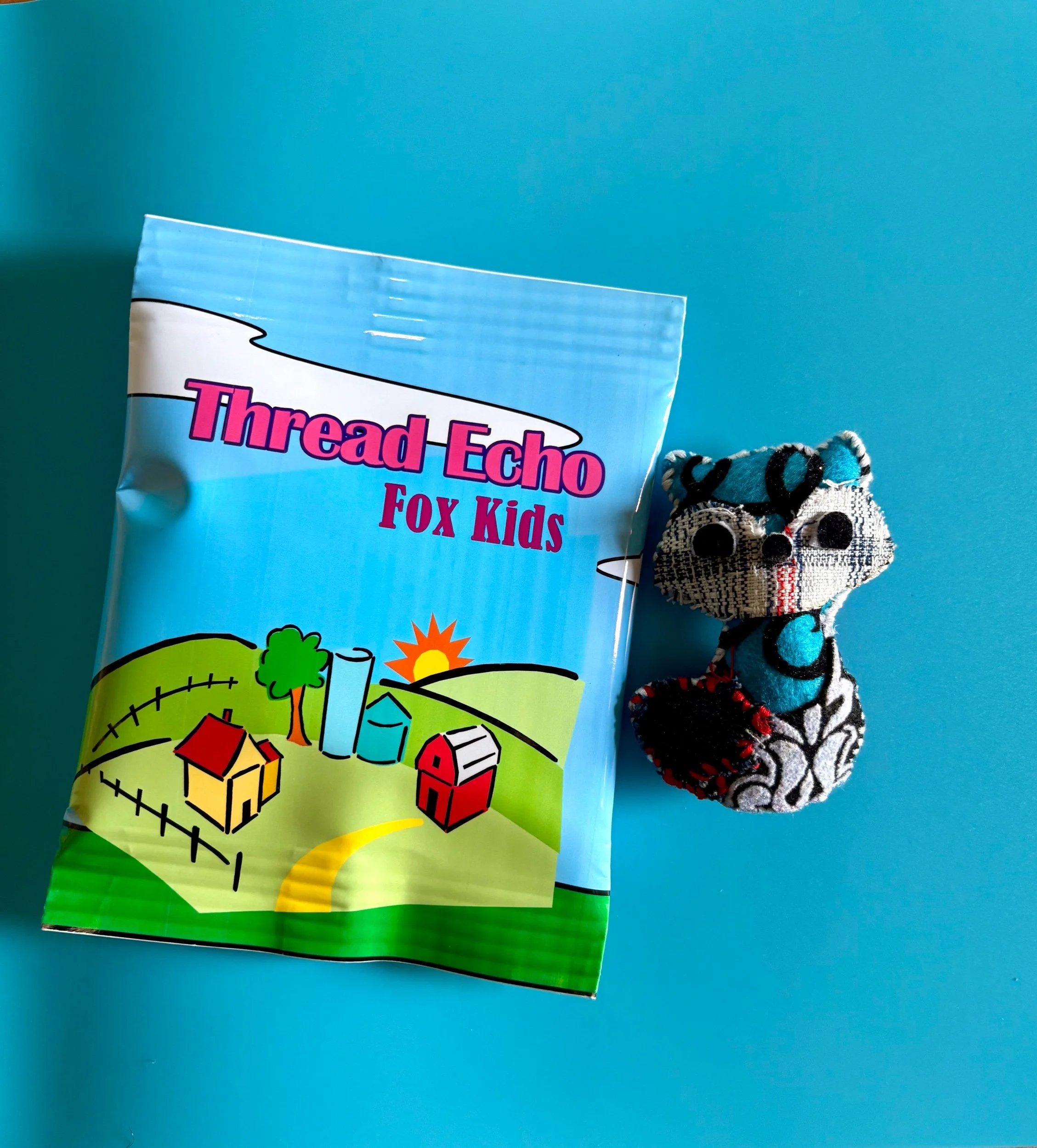 A packet labeled 'Thread Echo Fox Kids' with an illustration of a farm scene on a blue background, next to a small knitted fox figurine on a blue surface.