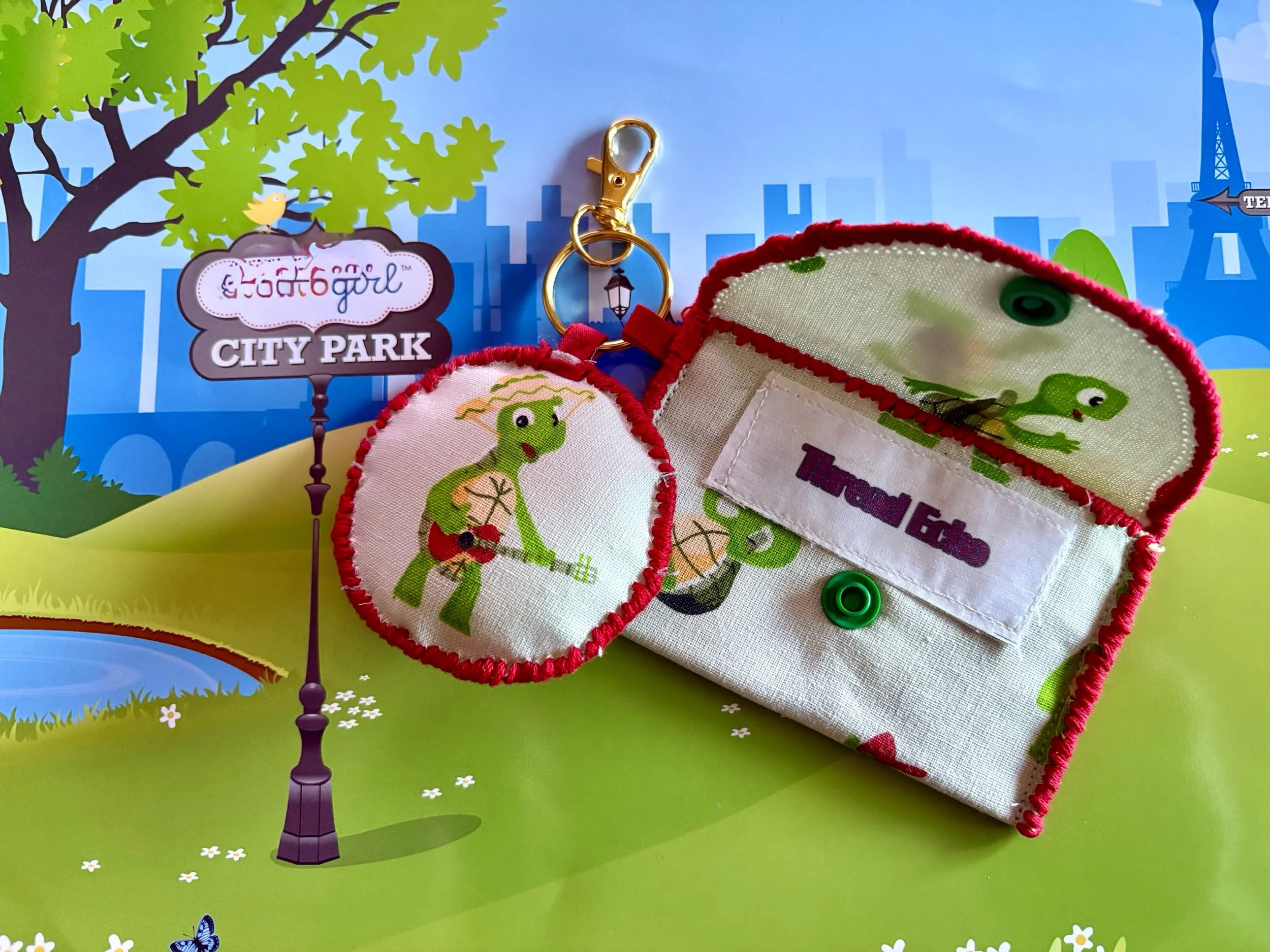 A keychain with a fabric circle featuring a green cartoon lizard playing a guitar, hanging next to a fabric pouch with a similar lizard print, on a colorful background of a cartoon park scene with trees, a pond, and a city skyline.