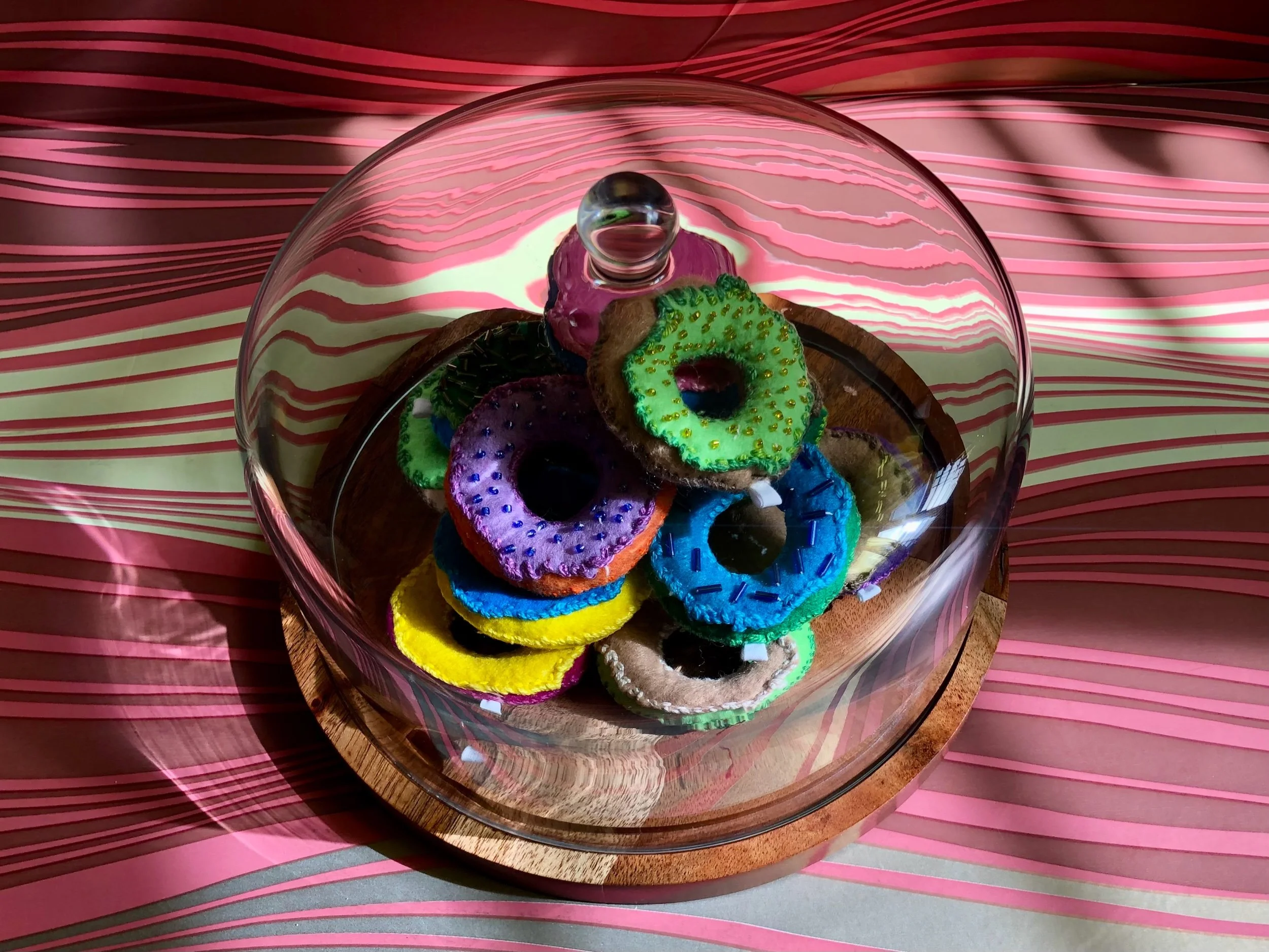 Colorful felt donuts inside a glass bowl placed on a wooden tray, on a pink and white striped surface.