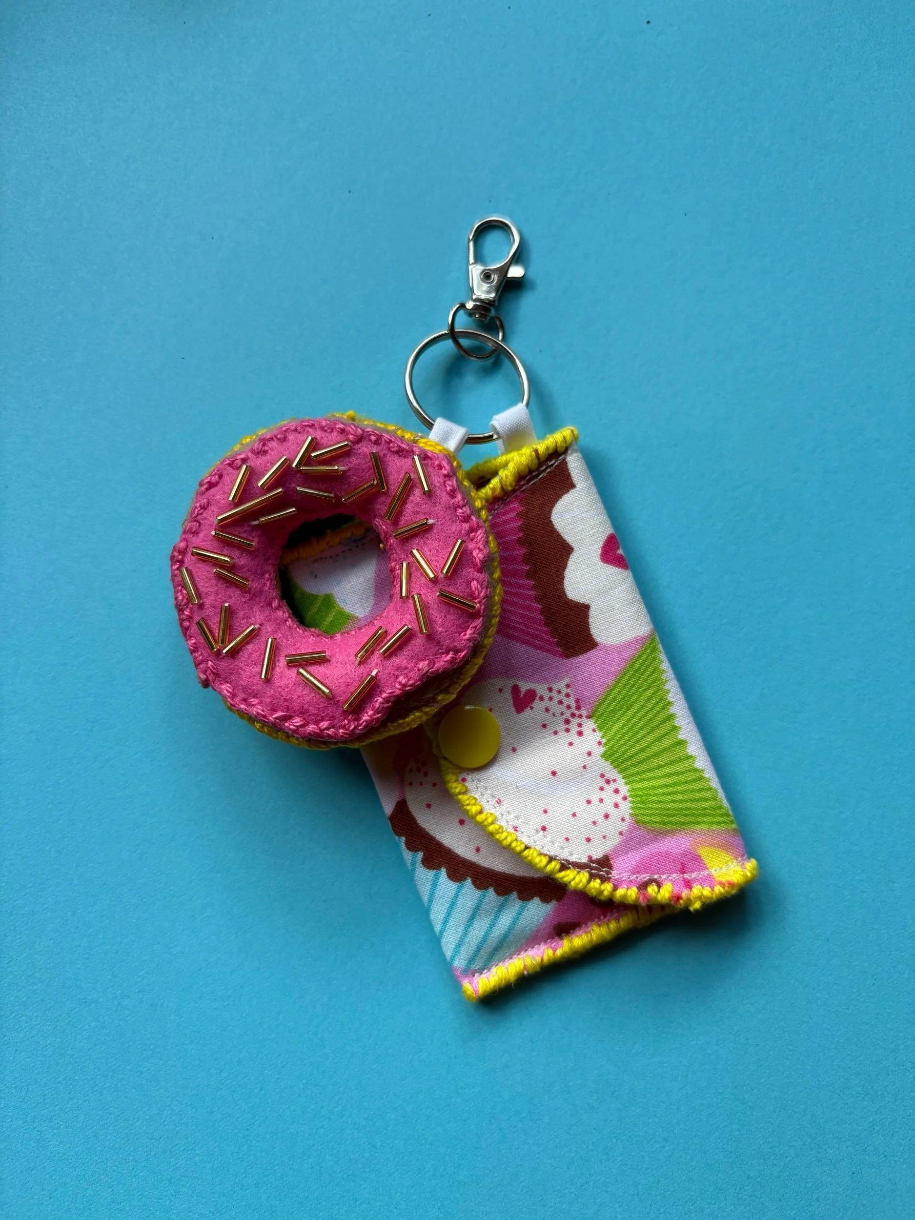 A plush pink donut keychain with gold sprinkles attached to a metal clip, and a fabric cupcake-themed keychain with a yellow button, connected to a metal keyring, on a blue background.