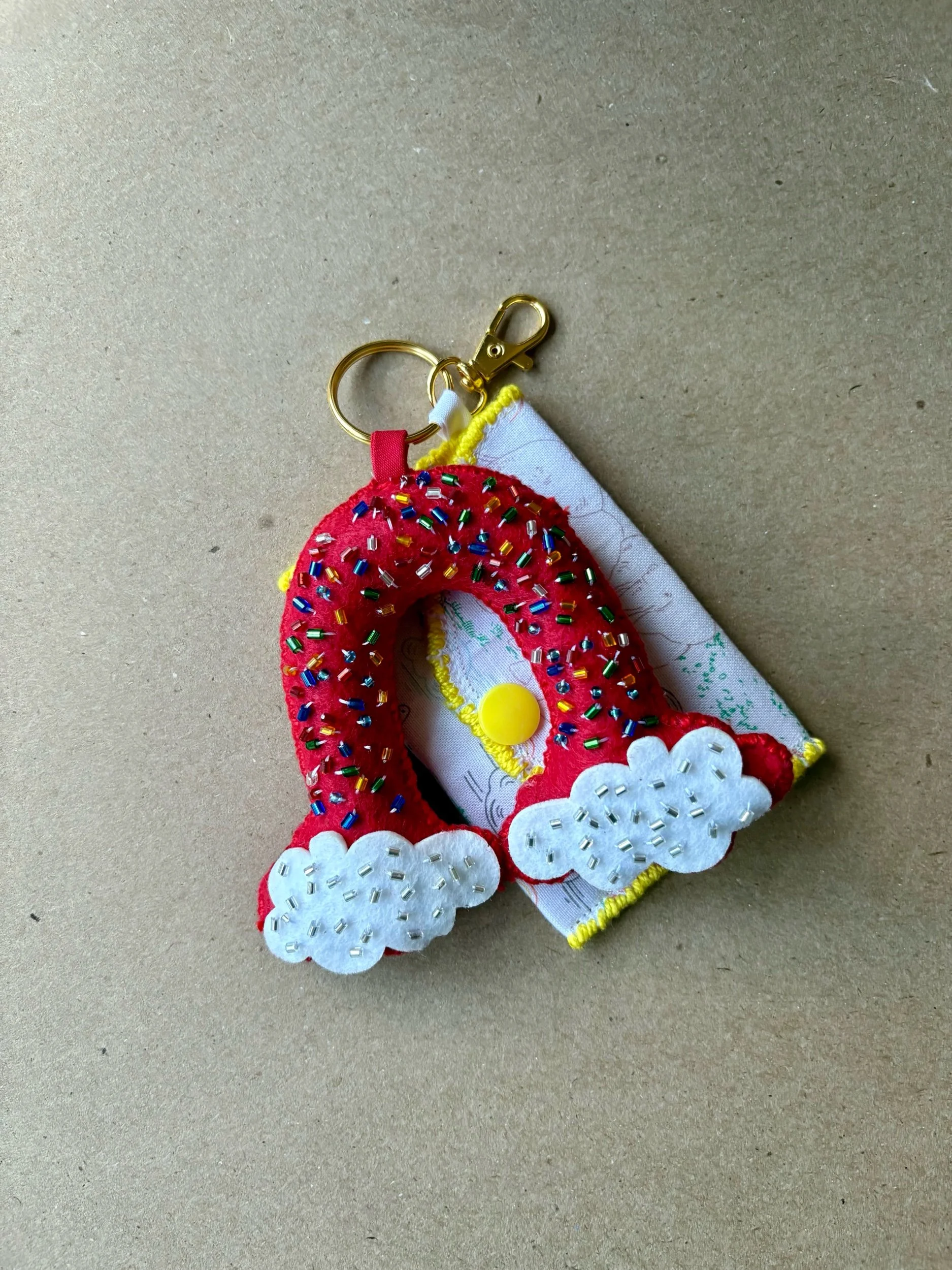 A fabric keychain shaped like a rainbow with white clouds, embroidered with beads, attached to a keyring and a small cloth piece with a yellow button.