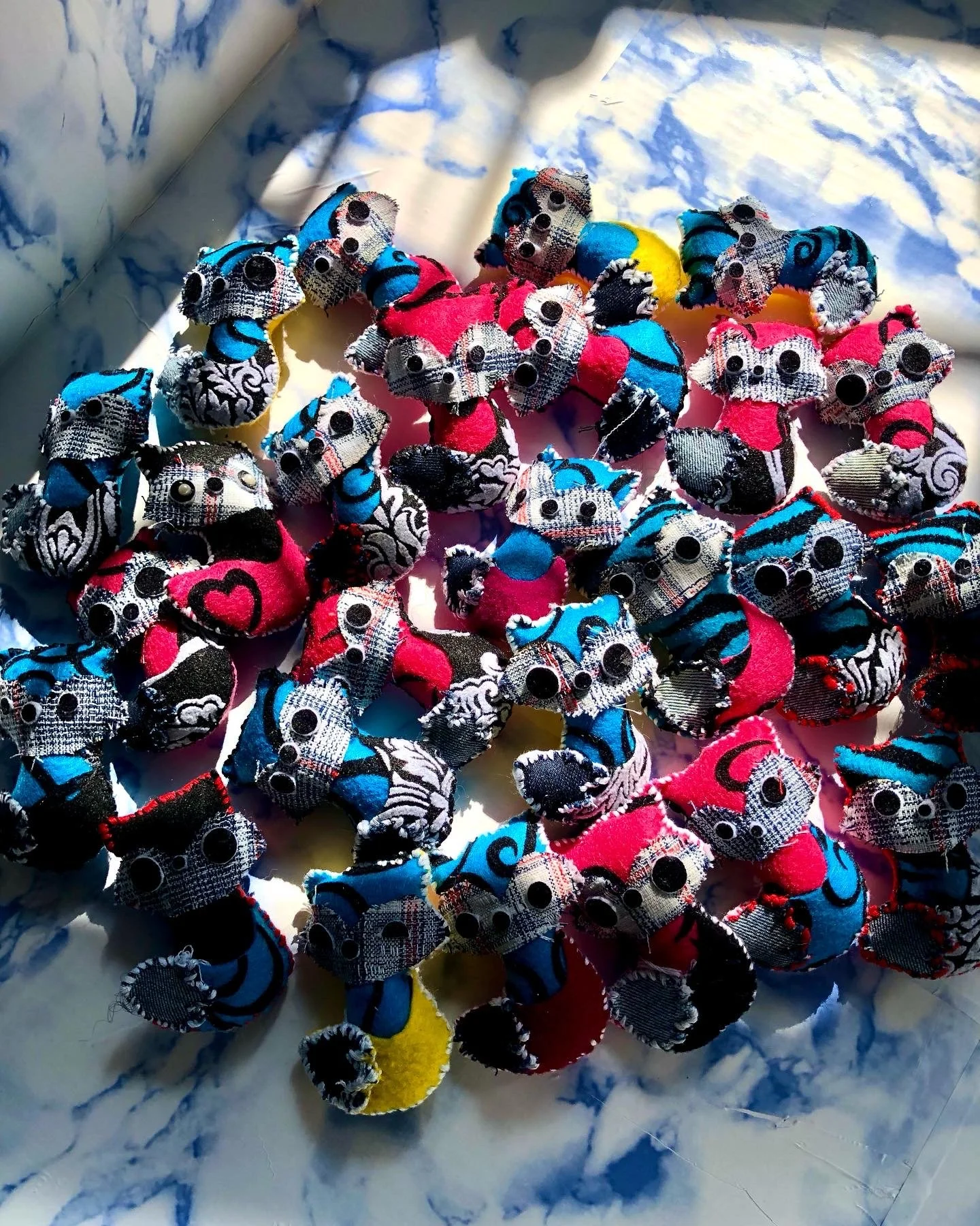 Collection of small fabric stuffed toys with googly eyes, arranged on a marble surface with sunlight casting shadows.