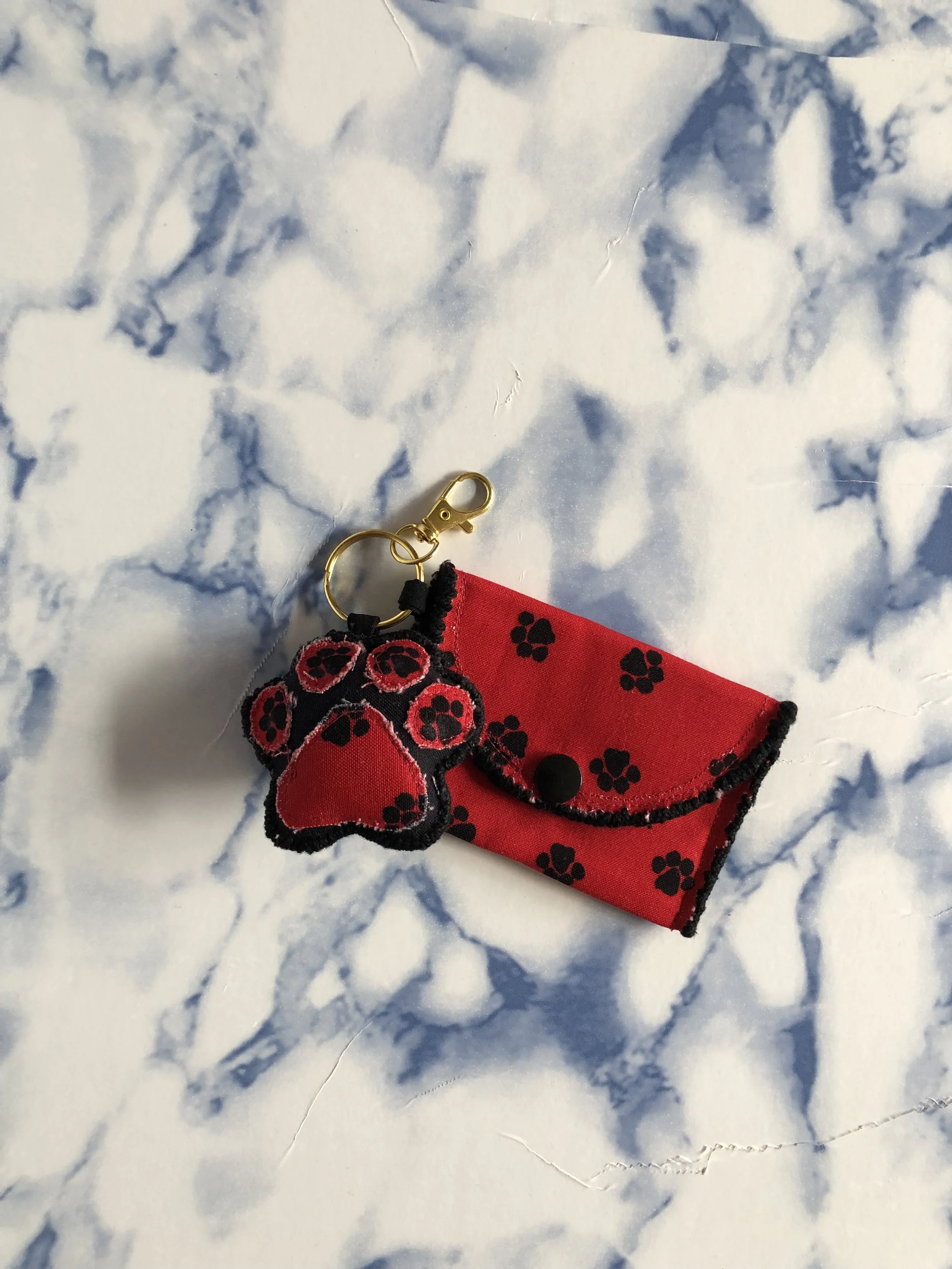 Red fabric keychain with black paw prints, black trim, and a paw-shaped attached charm, placed on a white and blue marble background.