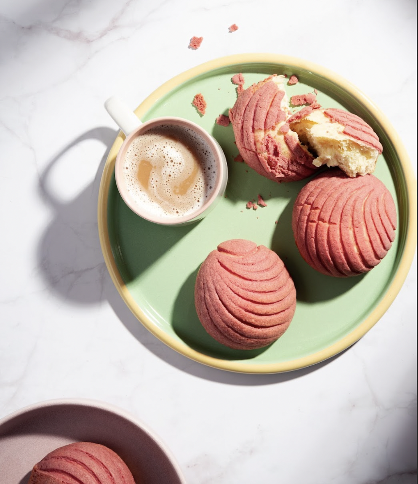 A green tray with three pink cookies and a cup of frothy coffee on a white marble surface. One cookie is broken showing a light interior.