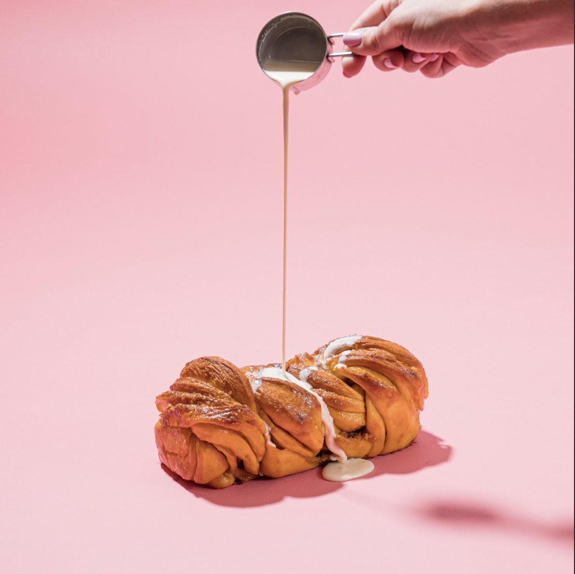 A hand pouring syrup over a twisted pastry on a pink background.