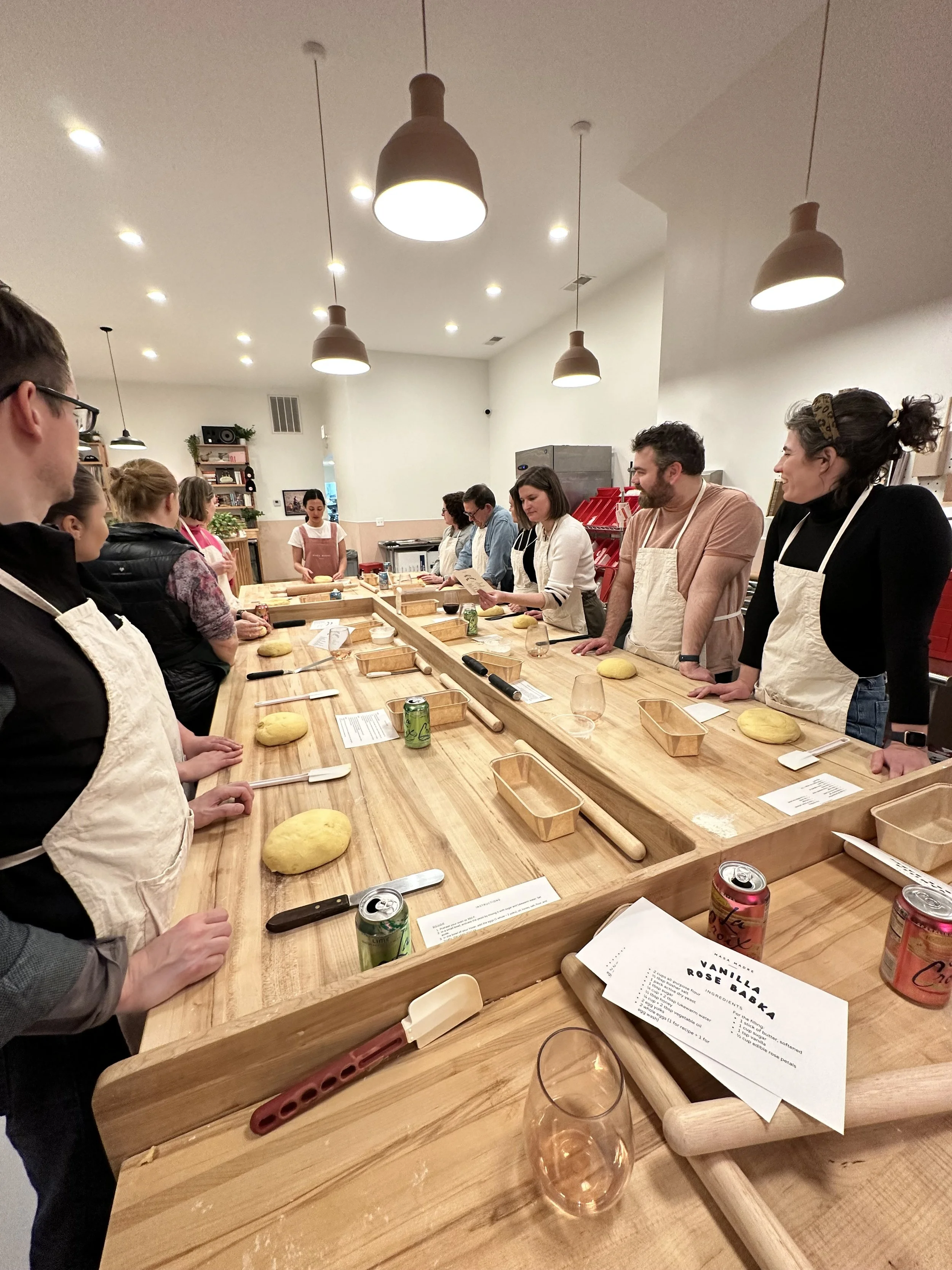 Group of people participating in a baking class around a wooden table with dough, utensils, and instructions, in a well-lit room.