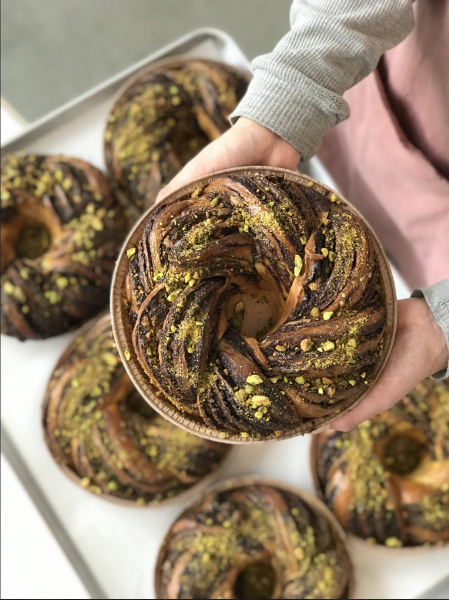 Hand holding a chocolate and pistachio babka pastry with chocolate swirls and chopped pistachios on top, with more babka pastries in the background.