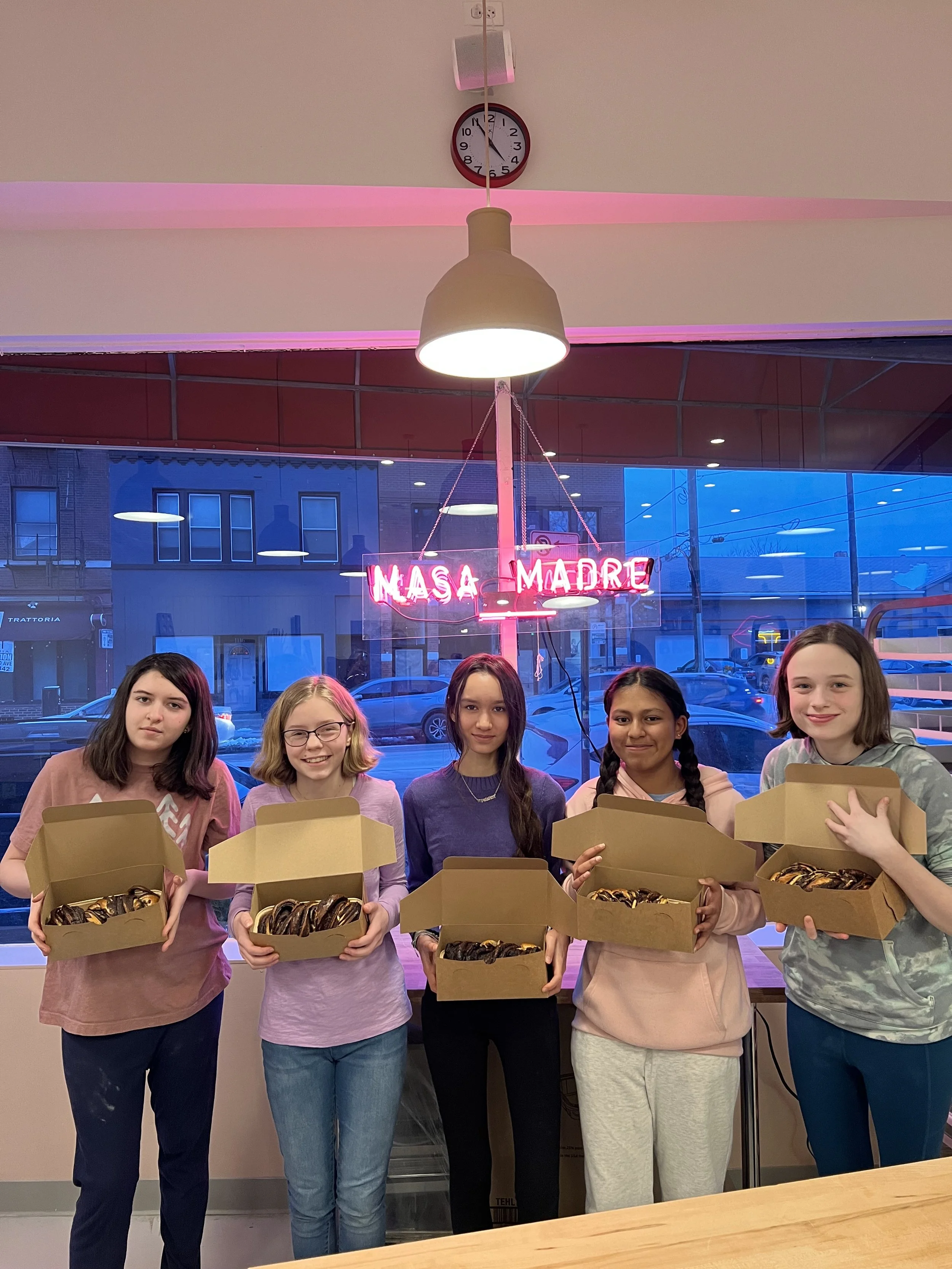 Five young girls standing in a row holding boxes of donuts in a cafe with neon sign reading 'NASA MADRE' in the background.