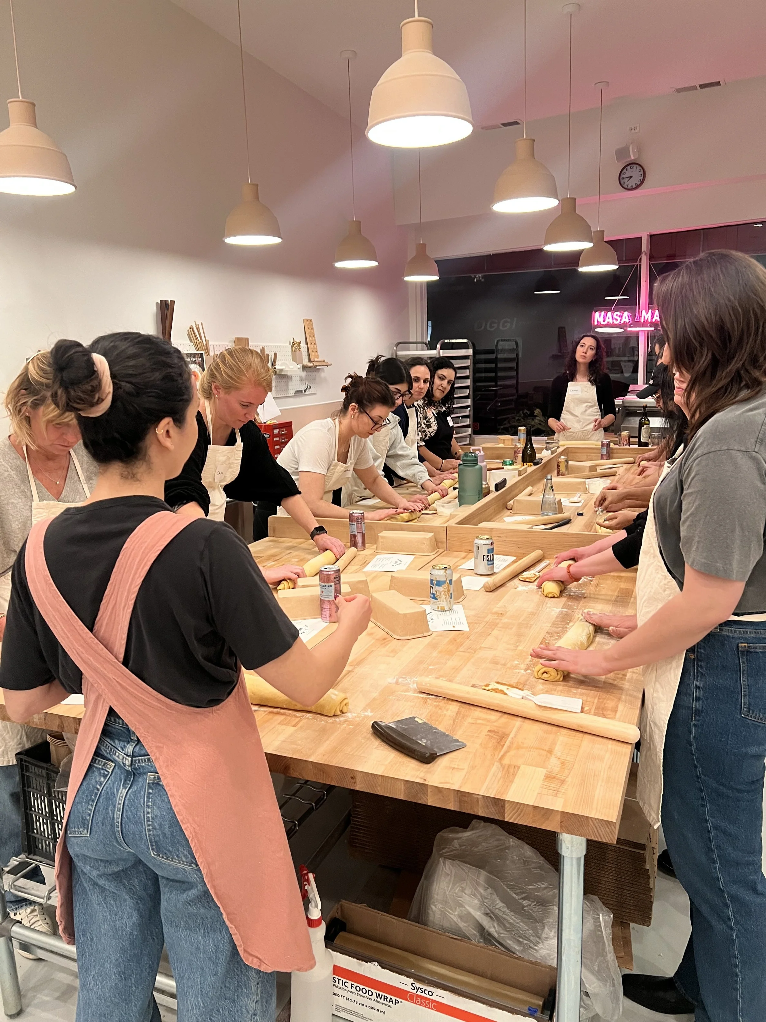 A group of women participating in a baking class, working with dough and rolling pins around a large wooden table in a modern kitchen studio.