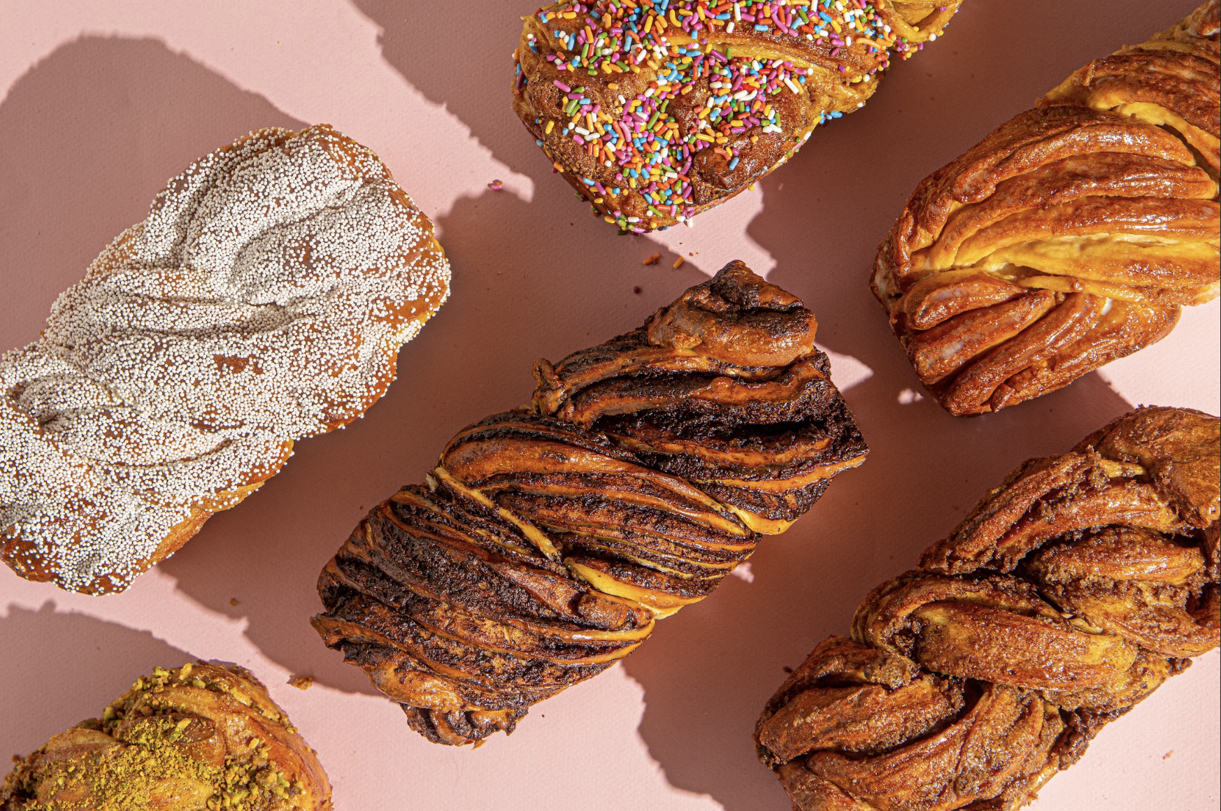 A variety of colorful pastries with different toppings, including sprinkles, sugar pearls, and crushed pistachios, on a pink background.