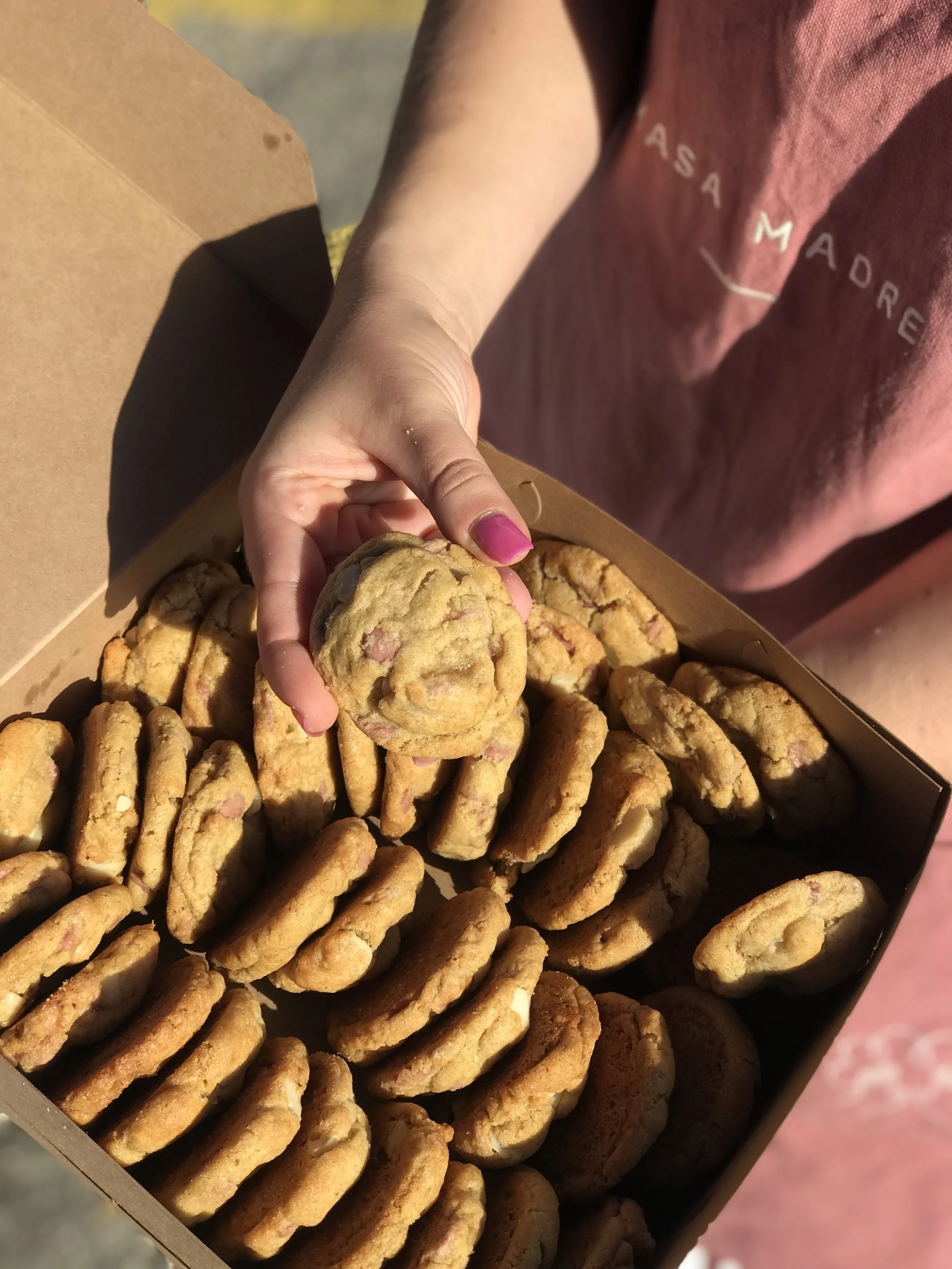 Person holding a cookie over a box of assorted cookies outdoors.