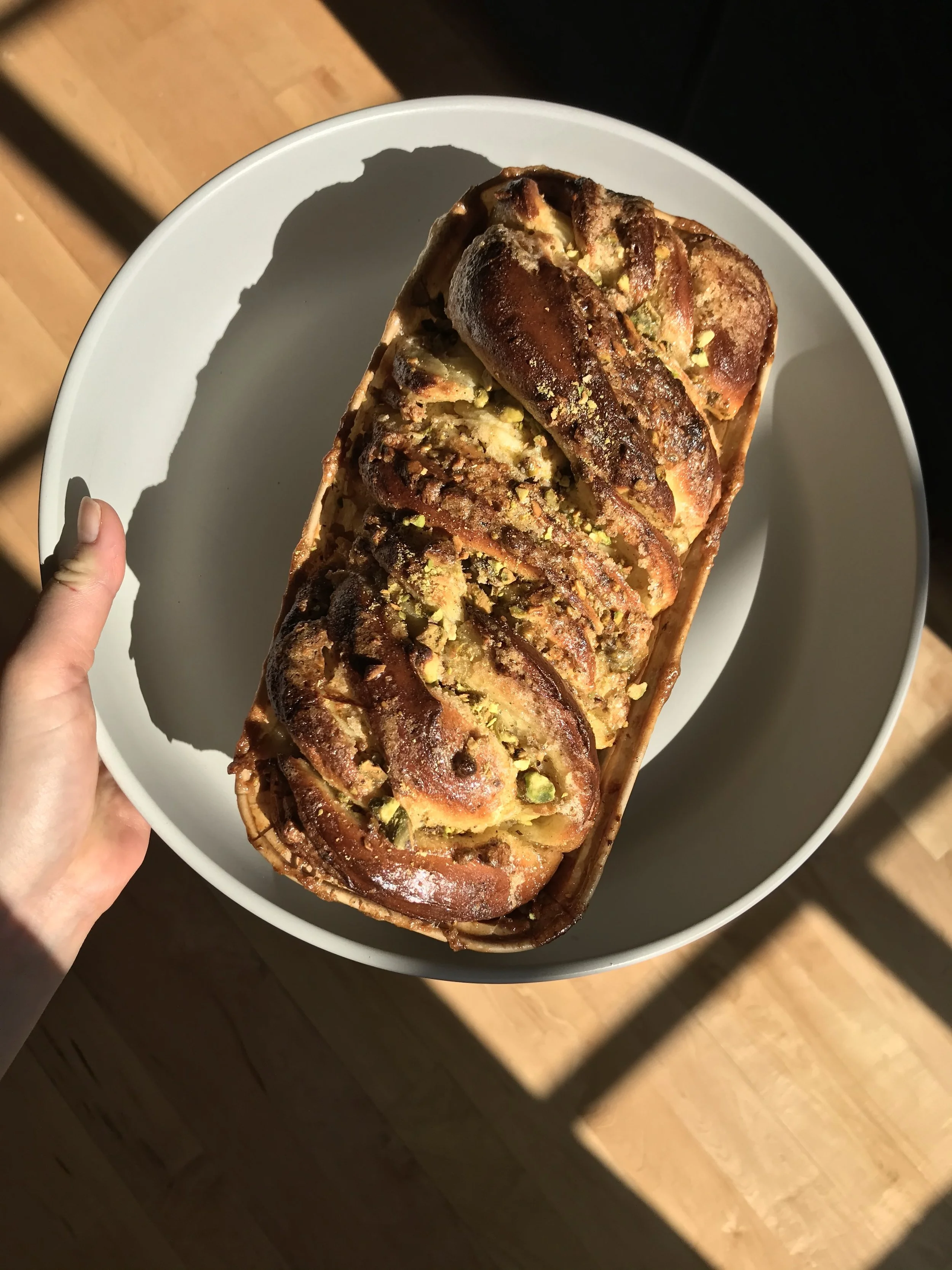 Top view of a freshly baked pistachio and chocolate babka on a white plate, held with a hand. The babka has a twisted shape with a golden-brown crust and visible green pistachios.