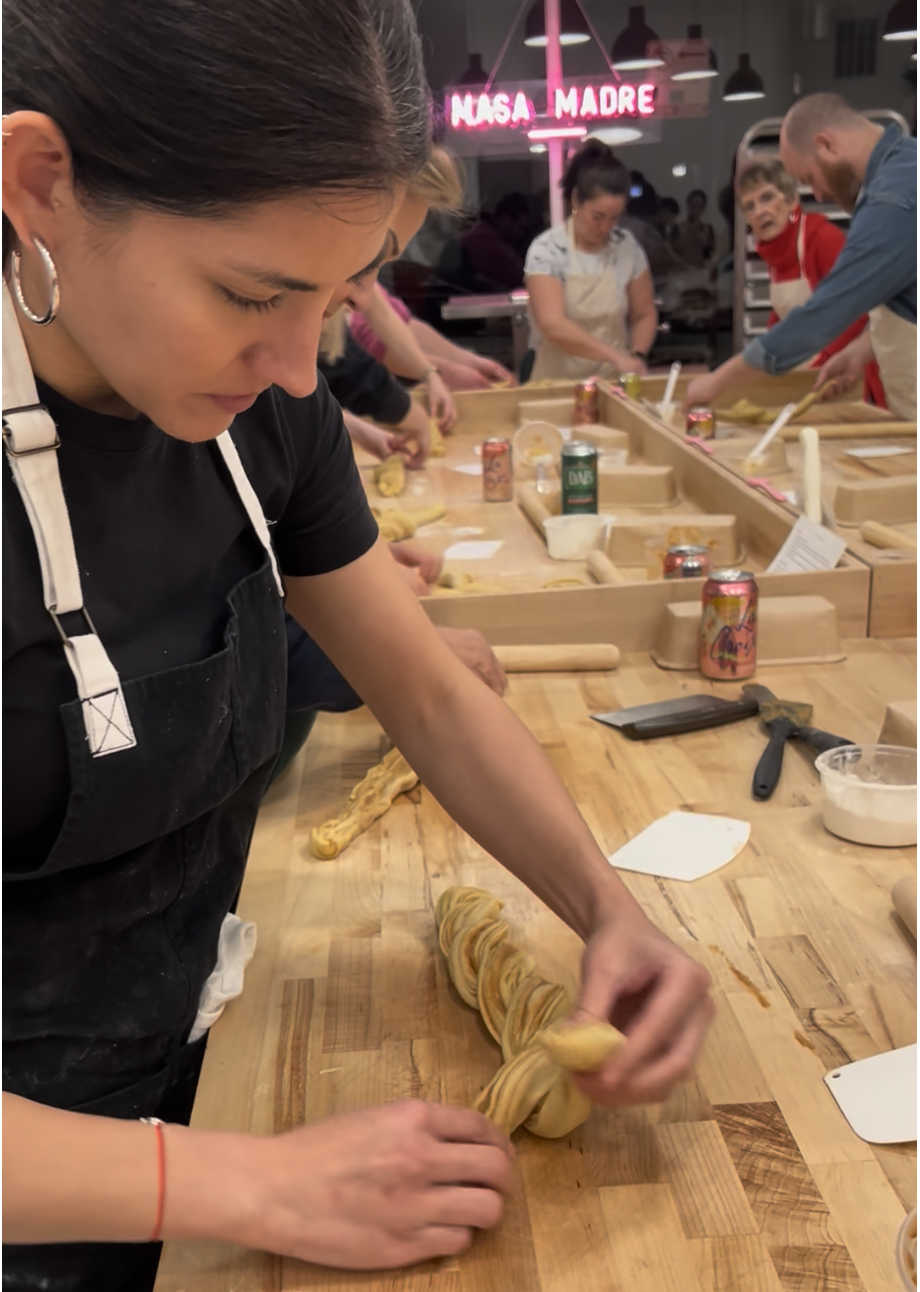 A woman working on twisted pasta dough at a wooden table in a cooking class, with other participants in the background and a pink neon sign that reads 'NASA MADRE'.