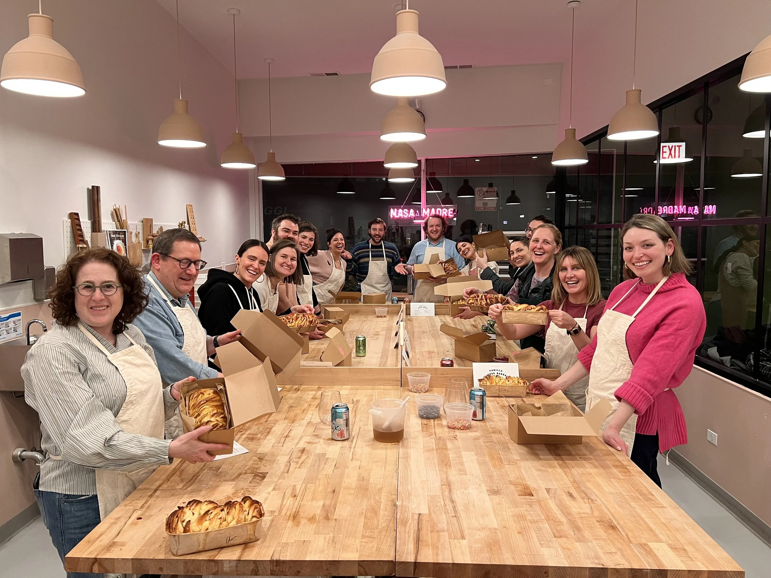 Group of people gathered around a large wooden table in a kitchen or bakery, holding open boxes of baked goods with smiles. The room has modern lighting, large windows, and a neon sign in the background.
