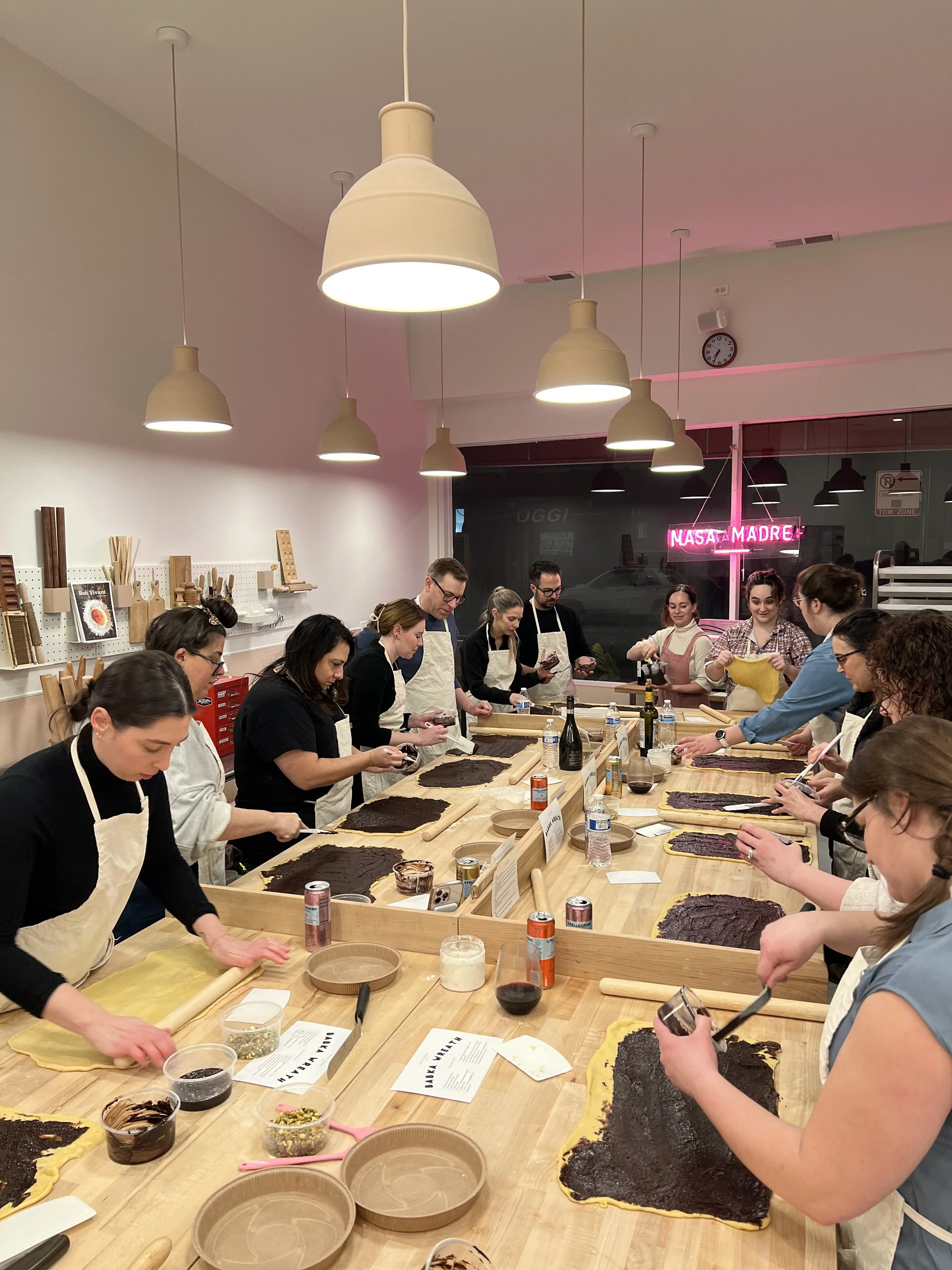 Group of people participating in a baking or cooking class, rolling out dough on tables with various ingredients and utensils, in a well-lit room with modern decor and a neon sign outside the window.