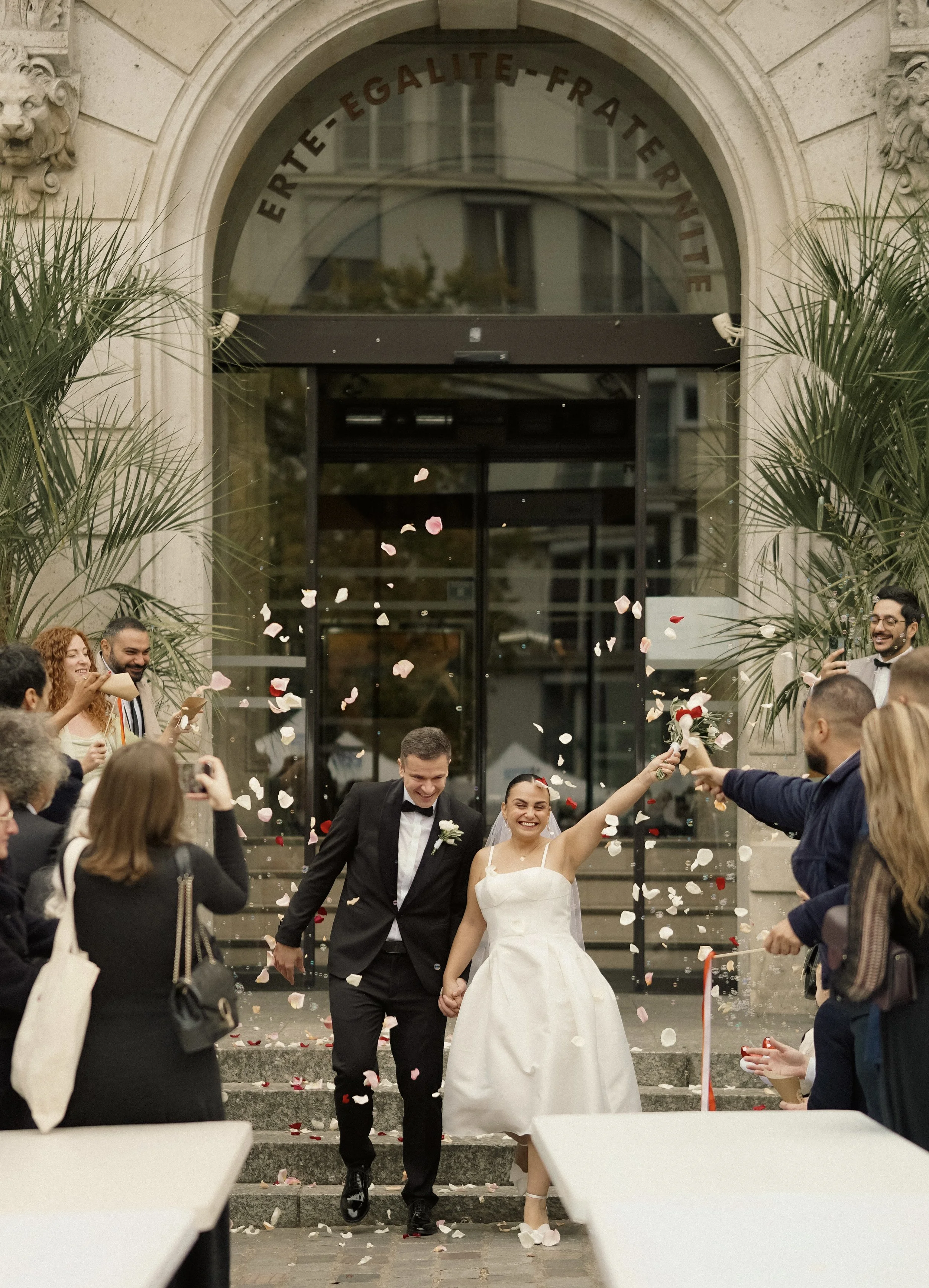 A newly married couple walking out of a building, holding hands, being celebrated by friends and family throwing flower petals.