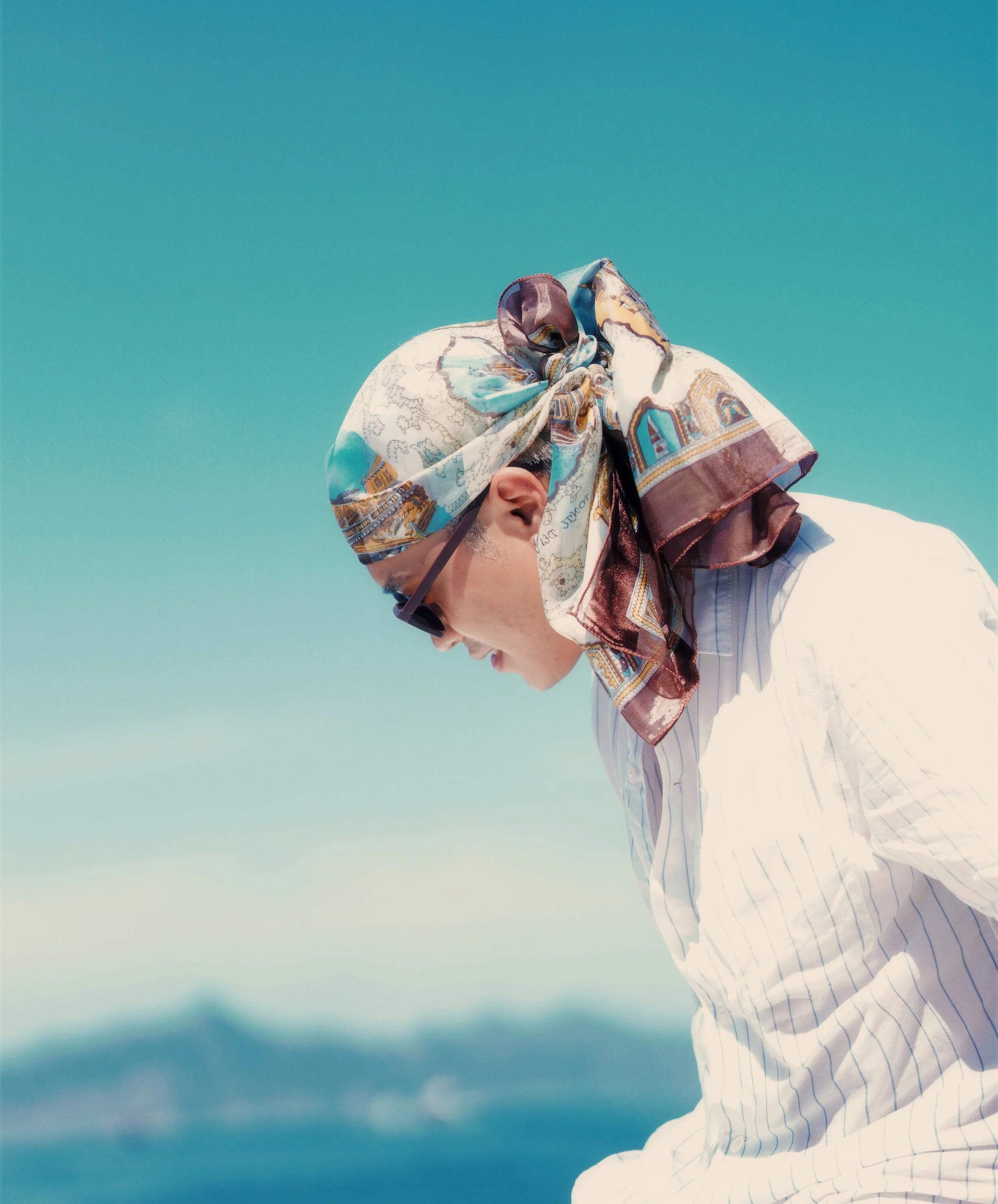 Person wearing a patterned headscarf and sunglasses, looking downward against a clear blue sky.