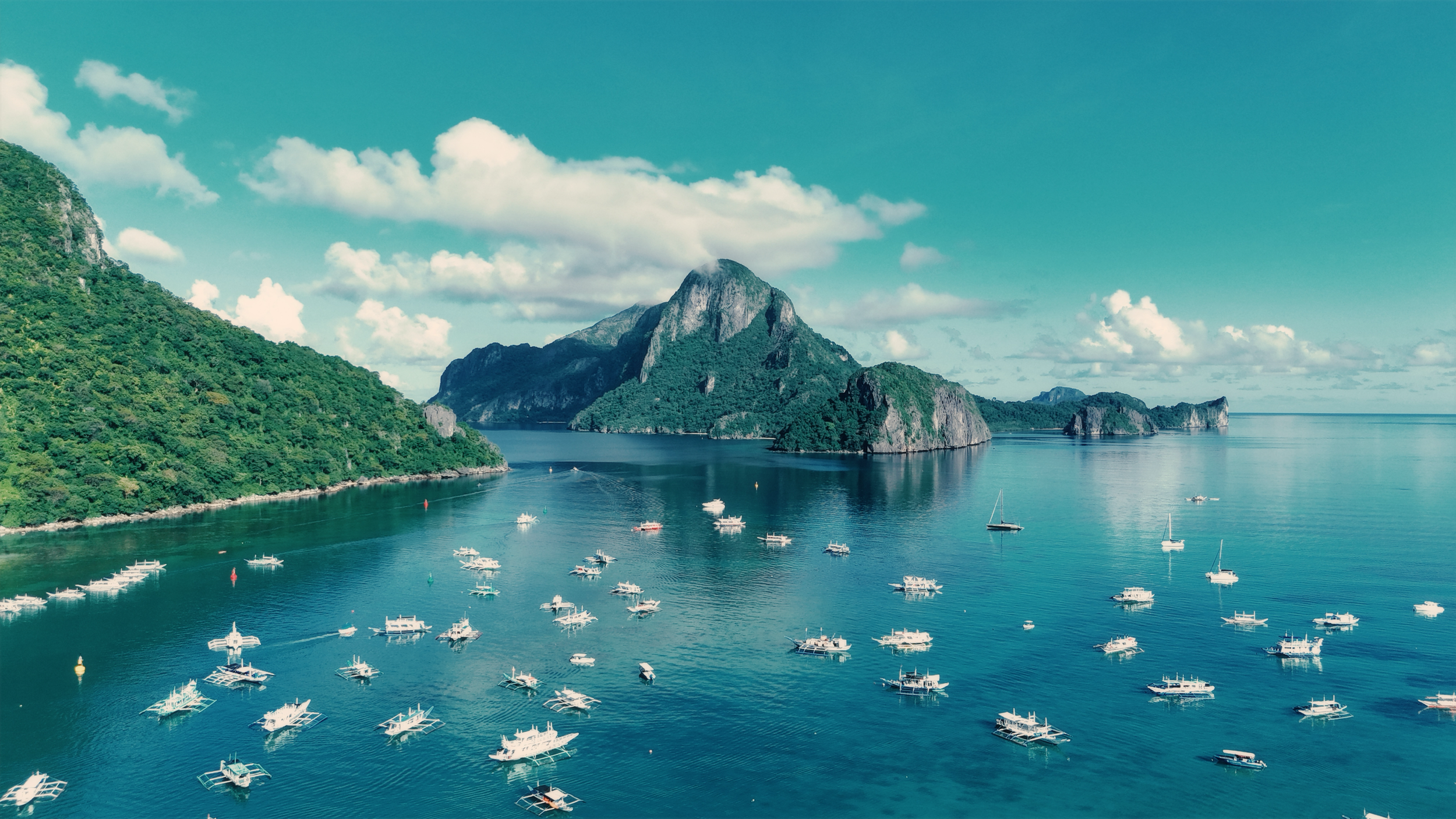 Tropical bay with numerous boats, surrounded by lush green islands and mountains, under a partly cloudy sky.
