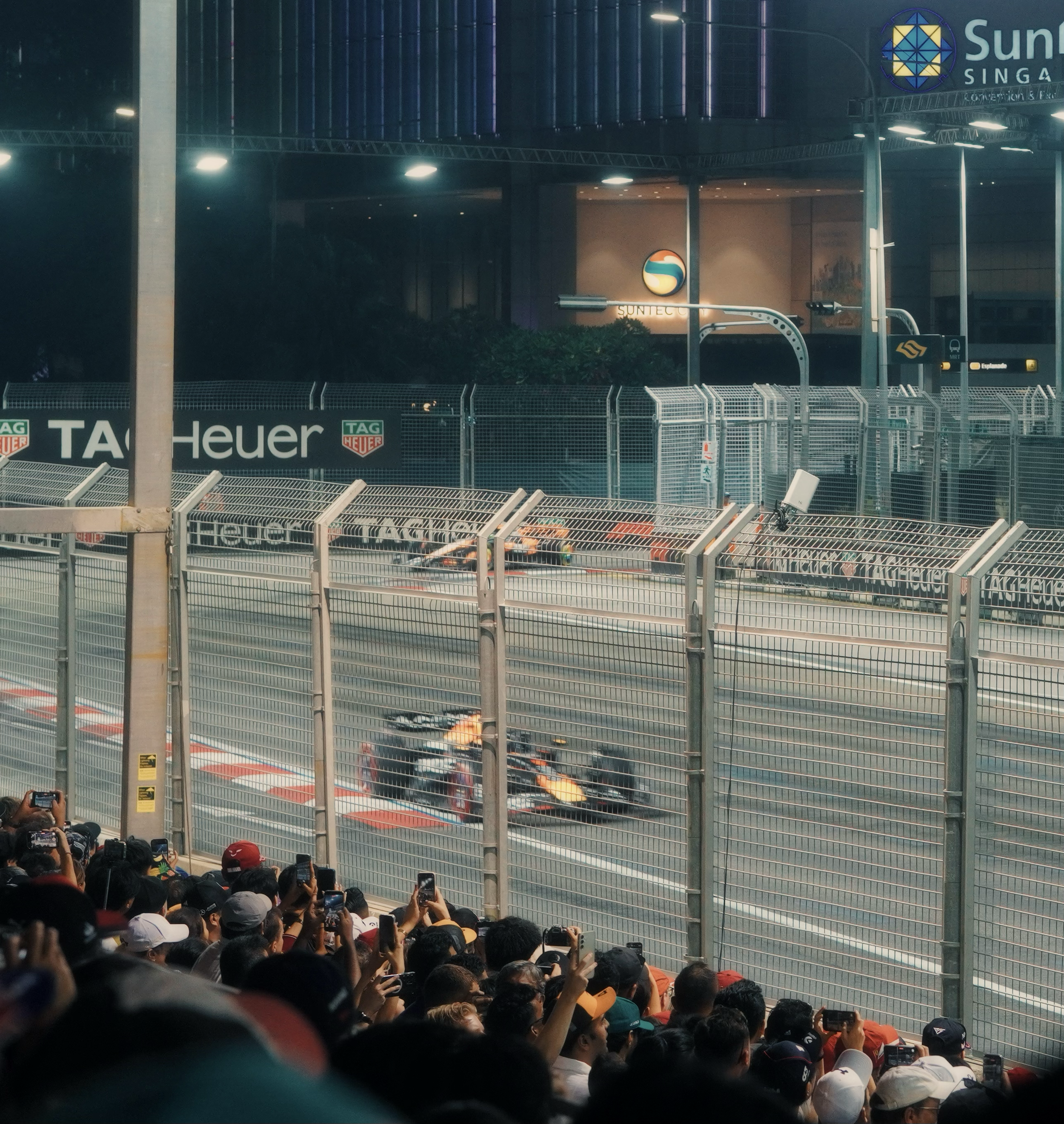 Formula 1 race cars speeding on a street circuit at night, viewed from the crowd behind a fence, with spectators capturing the moment on their phones.