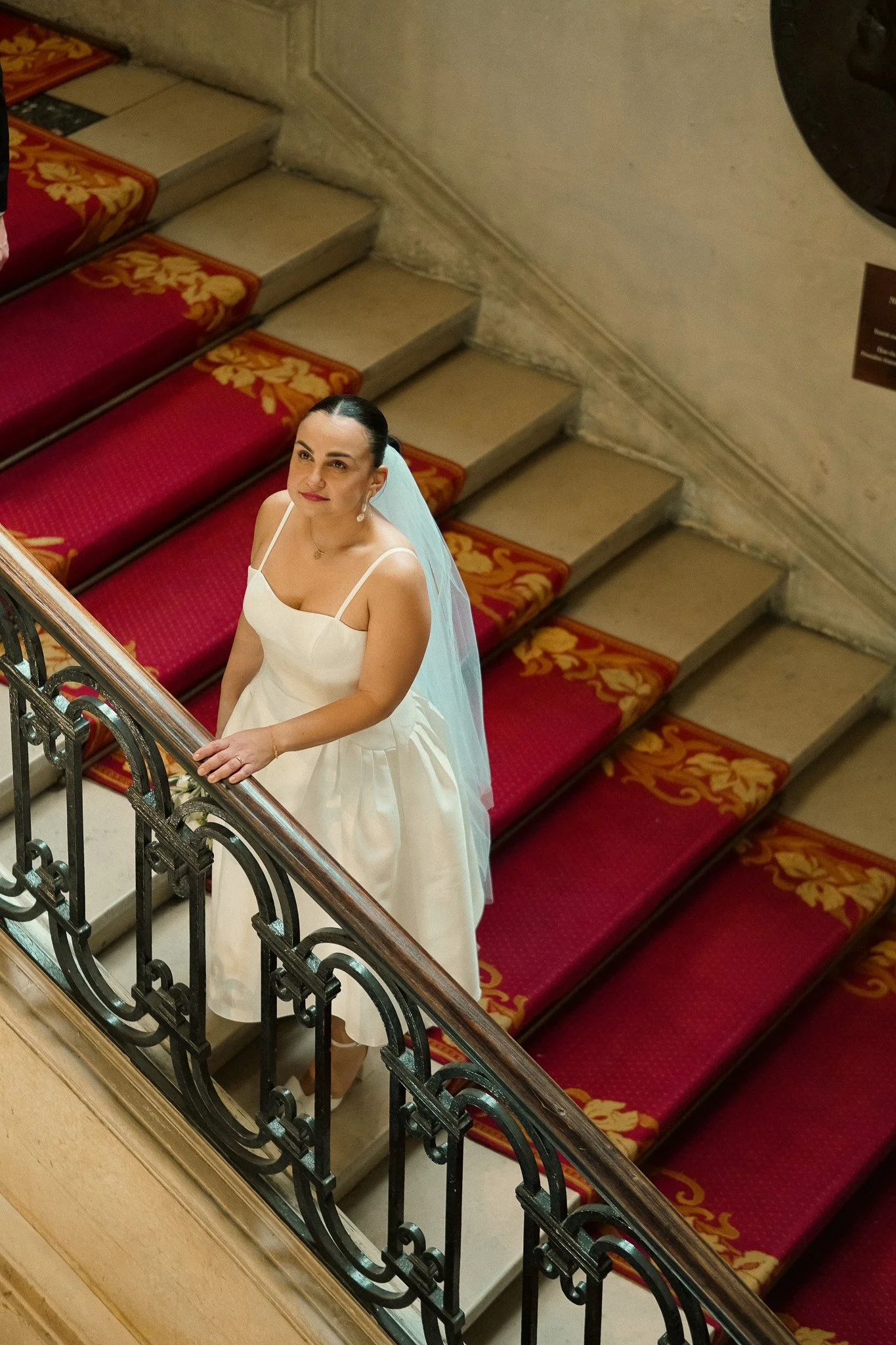 A bride in a white wedding dress standing on a staircase with red carpet, looking up at the camera.