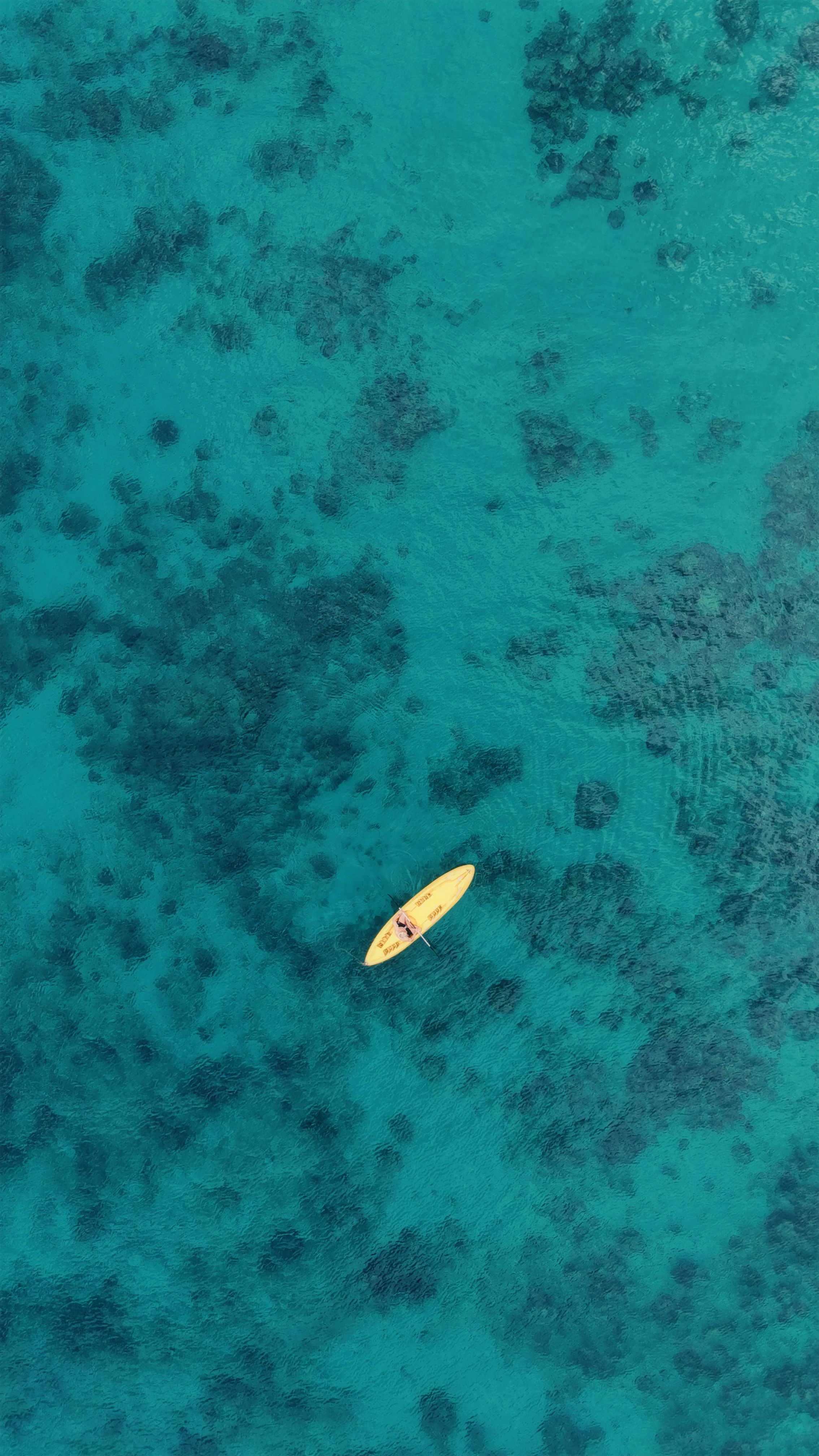 An aerial view of a person in a yellow kayak on clear, blue ocean water with visible underwater coral formations.