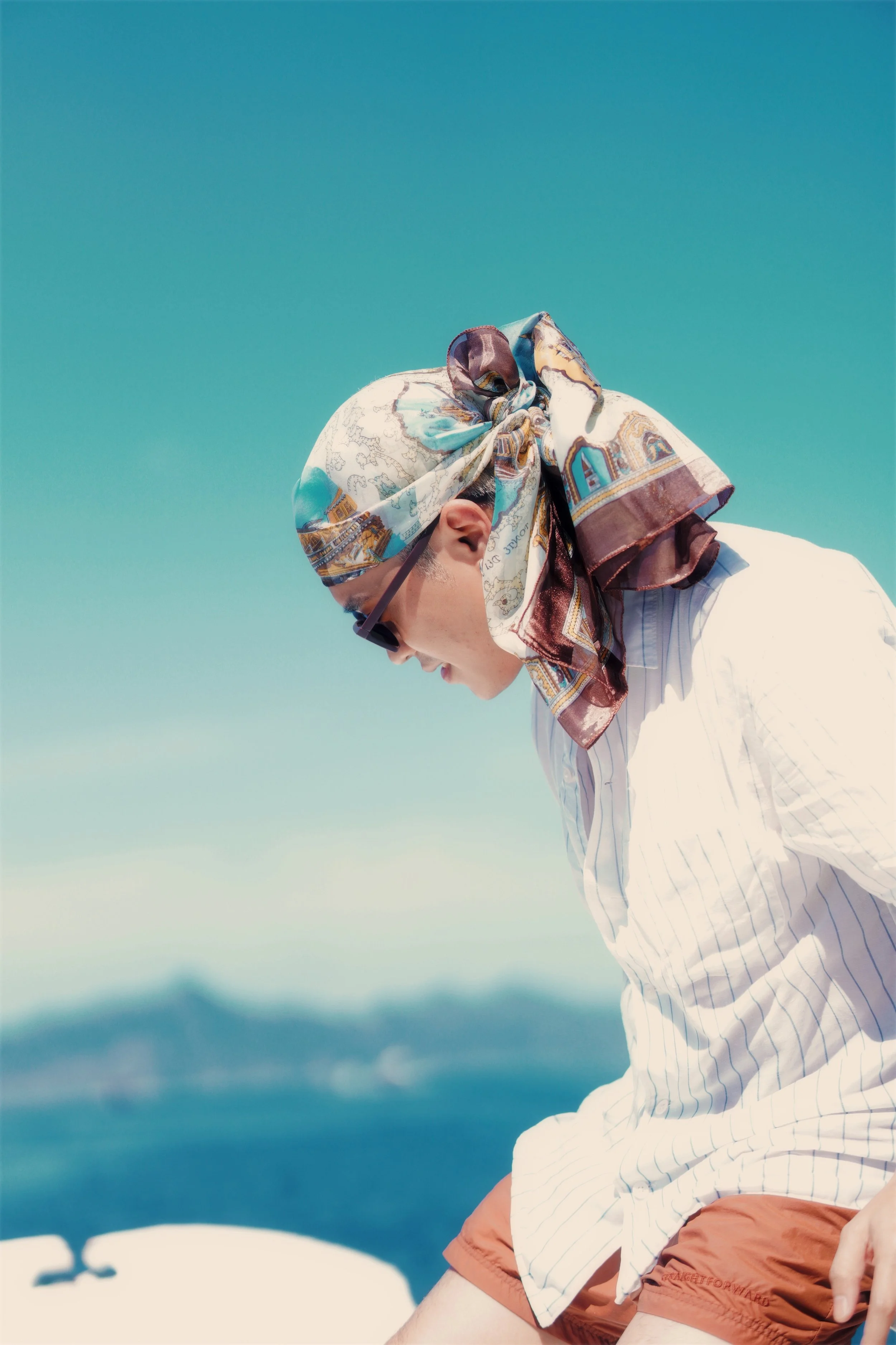 A man wearing a patterned scarf, sunglasses, a white striped shirt, and orange shorts, sitting outdoors against a clear blue sky with a blurred landscape in the background.