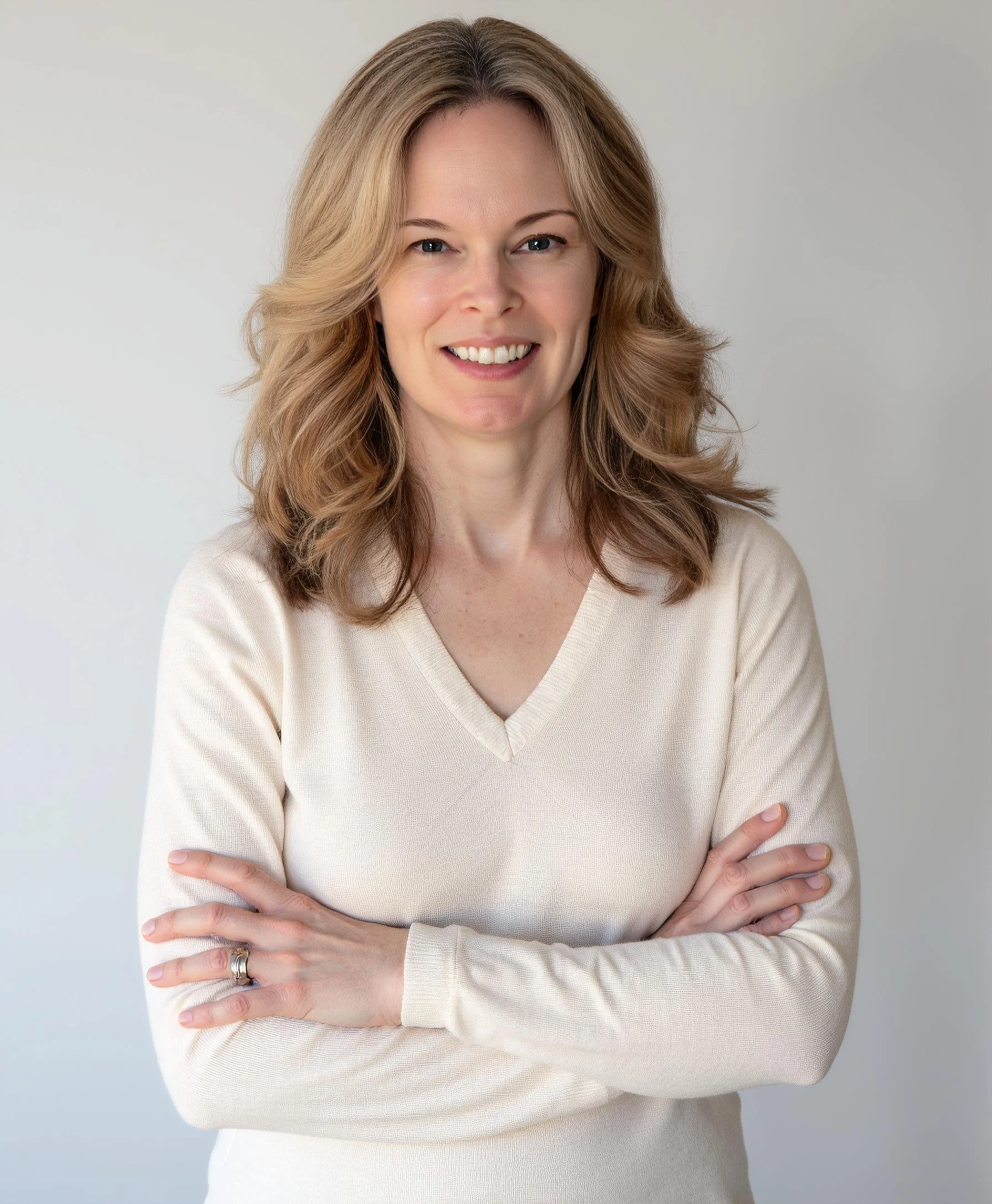 A headshot of Amy Woodman with wavy blonde hair, smiling, wearing a cream-colored V-neck sweater, arms crossed, standing against a plain light background.