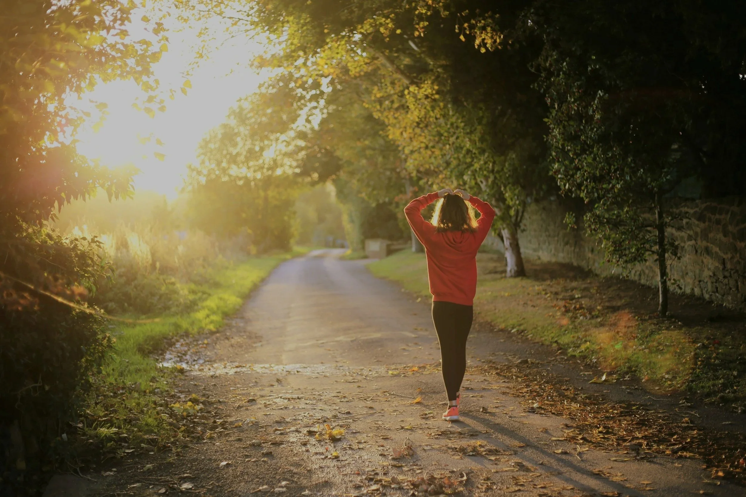 A woman jogging on a tree-lined path during sunset, with sunlight filtering through the leaves.