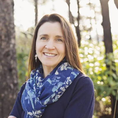 A woman with long brown hair smiling outdoors, wearing a blue floral scarf and dark clothing, in a forest setting with sunlight filtering through trees.