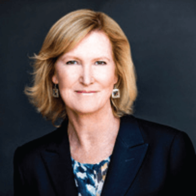 A woman with shoulder-length blonde hair wearing a dark blazer and patterned blouse, smiling against a dark background.