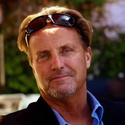 A middle-aged man with a beard and mustache, wearing sunglasses on his head, a blue shirt, and a black blazer, smiling outdoors with blurred greenery and pink flowers in the background.