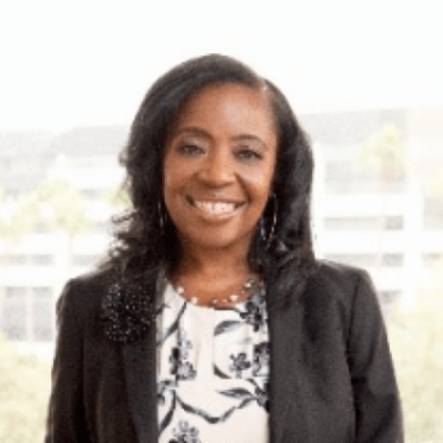 Professional woman with dark hair smiling, wearing a black blazer and a patterned blouse, standing outdoors with a cityscape in the background.