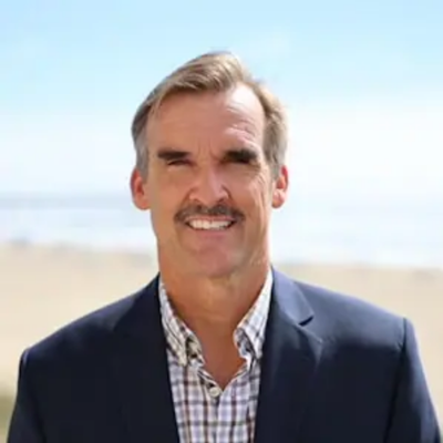 A middle-aged man with gray hair and a mustache smiling at the camera, standing outdoors on a beach with the ocean in the background.