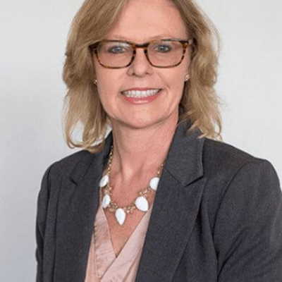 A woman with blonde hair and glasses, wearing a dark blazer and a beaded necklace, smiling at the camera.