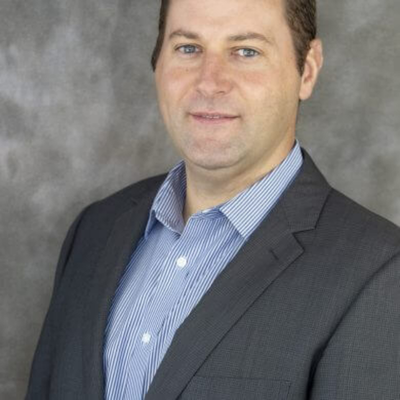 Professional headshot of a man in a suit and blue striped shirt against a gray background.