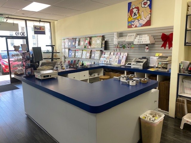 Inside a small retail store checkout counter with various merchandise, including greeting cards, gift wraps, and printers, with a window showing a FedEx sign and cars outside.