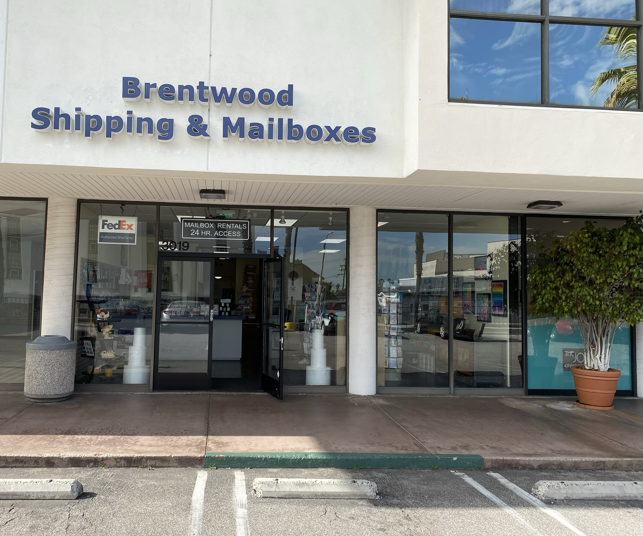 Front of a shipping and mailbox store called Brentwood Shipping & Mailboxes with a glass storefront, a sign indicating mailbox rentals with 24-hour access, and a parking lot in front.