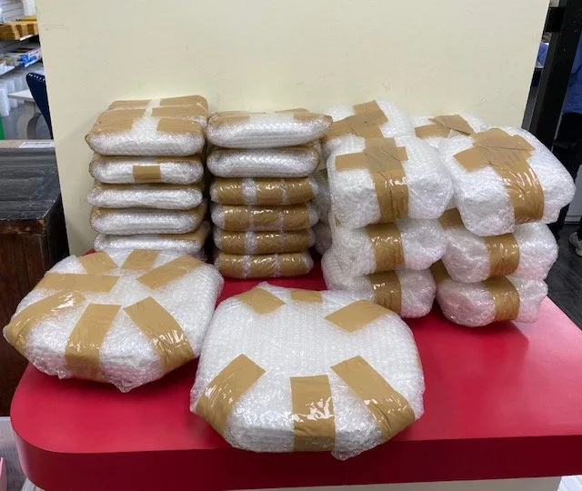 Multiple packages wrapped in bubble wrap and sealed with brown packing tape are stacked on a red table against a plain wall.