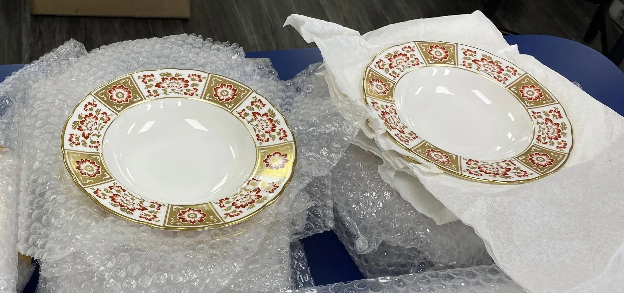 Two decorative plates with floral patterns and gold rims, wrapped in tissue paper and surrounded by bubble wrap.