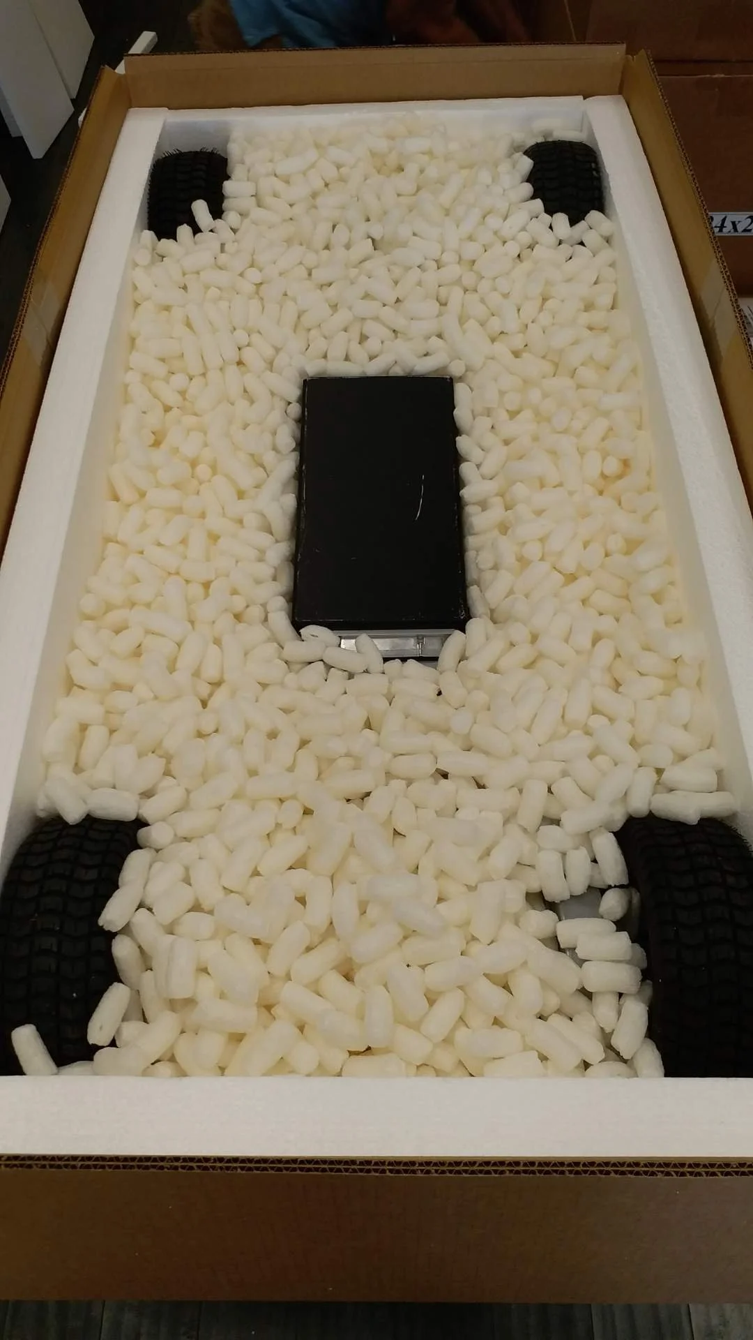 A cardboard box filled with white packing peanuts, with a black smartphone in the center and four black rubber tires at each corner.