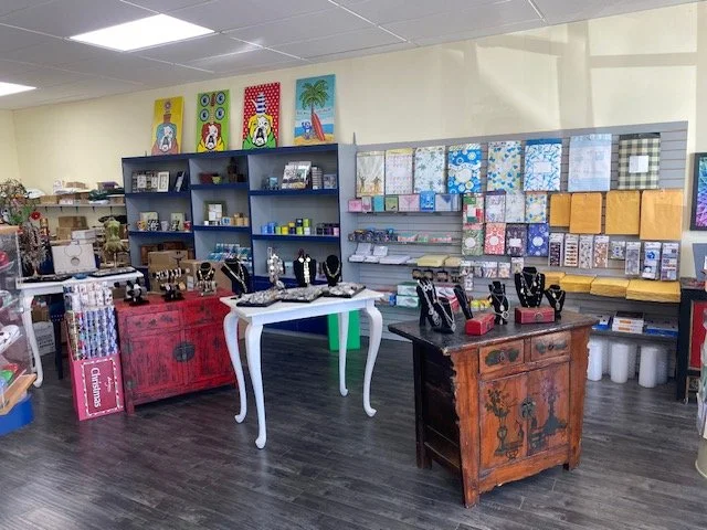 Display of jewelry on black bust stands on a white table and a wooden table in a gift shop, with shelves behind filled with stationery and colorful artwork on the walls.