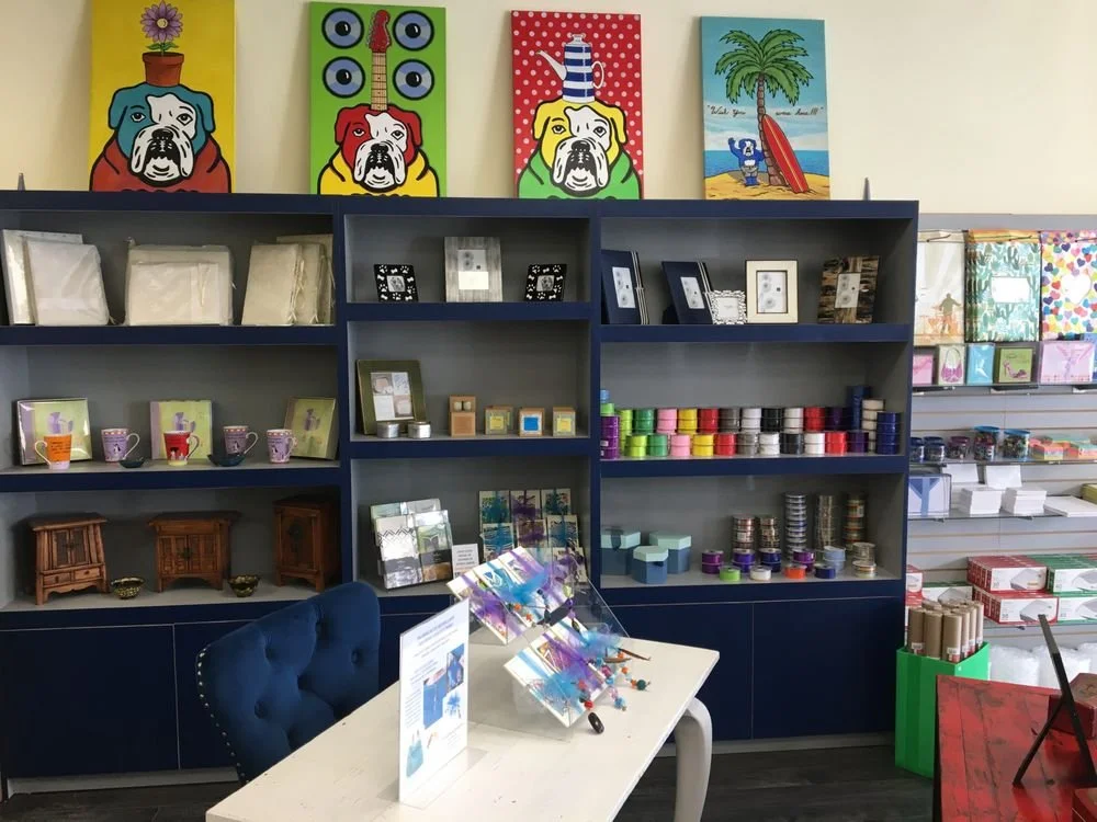 Interior of a store with blue shelving units displaying decorative items, picture frames, and art supplies. Colorful paintings of dogs with various backgrounds are hung above the shelves.