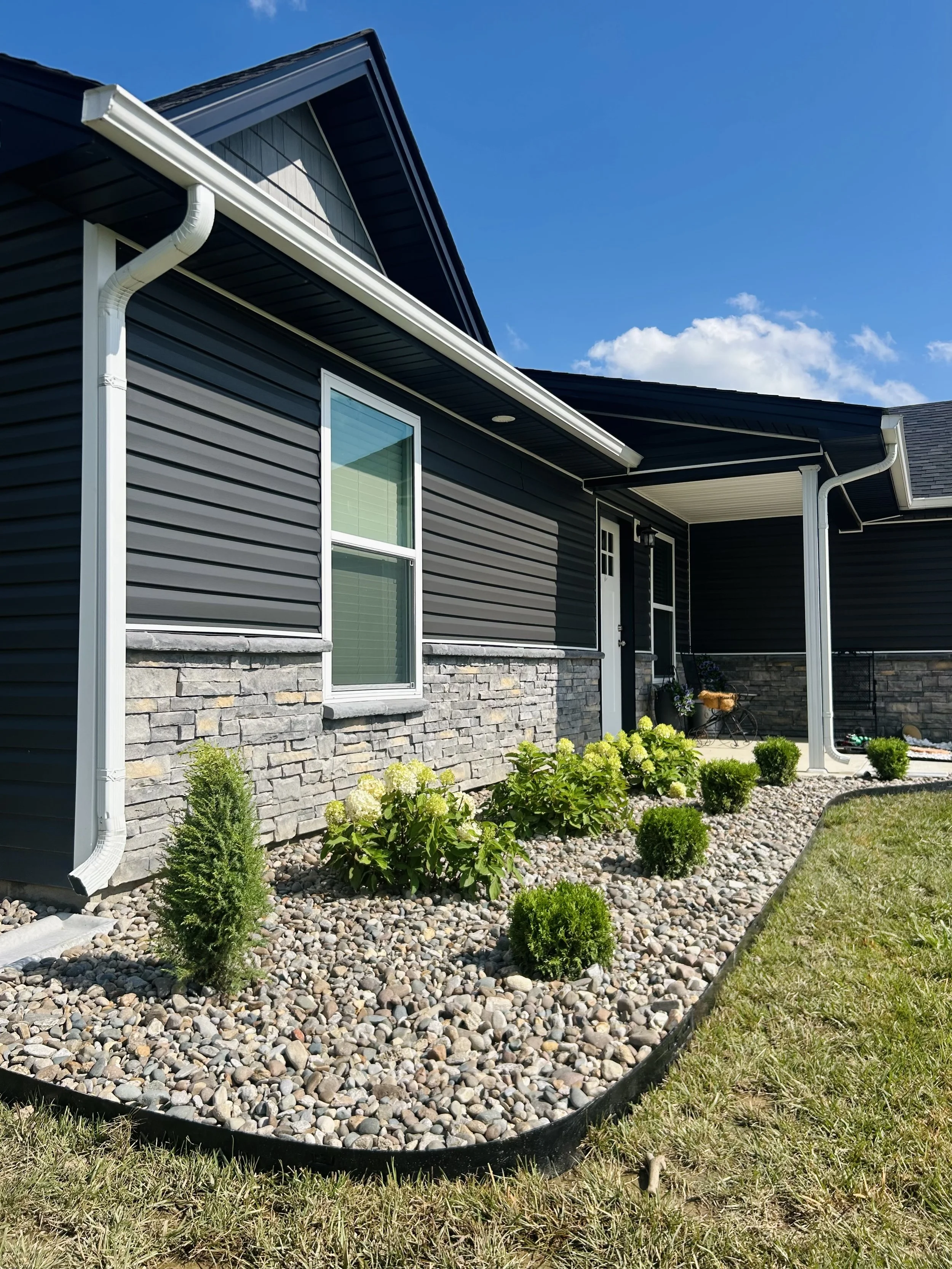 Front yard with small bushes, flowers, and gravel border in front of a modern house with dark siding, stone detail, and white trim under a blue sky.