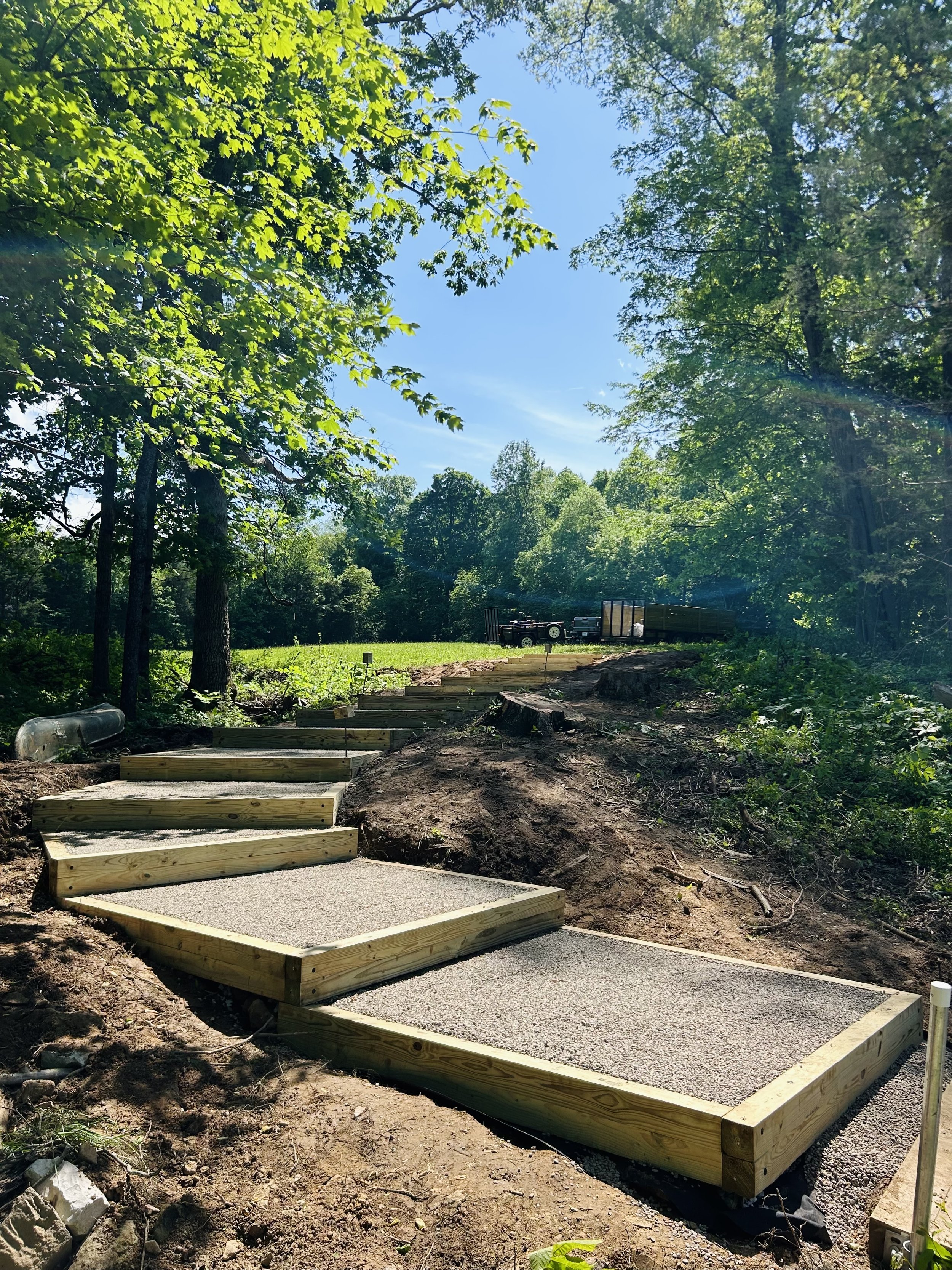 A winding pathway under construction in a wooded area, with sections of wooden framing and gravel, surrounded by green trees and a blue sky.